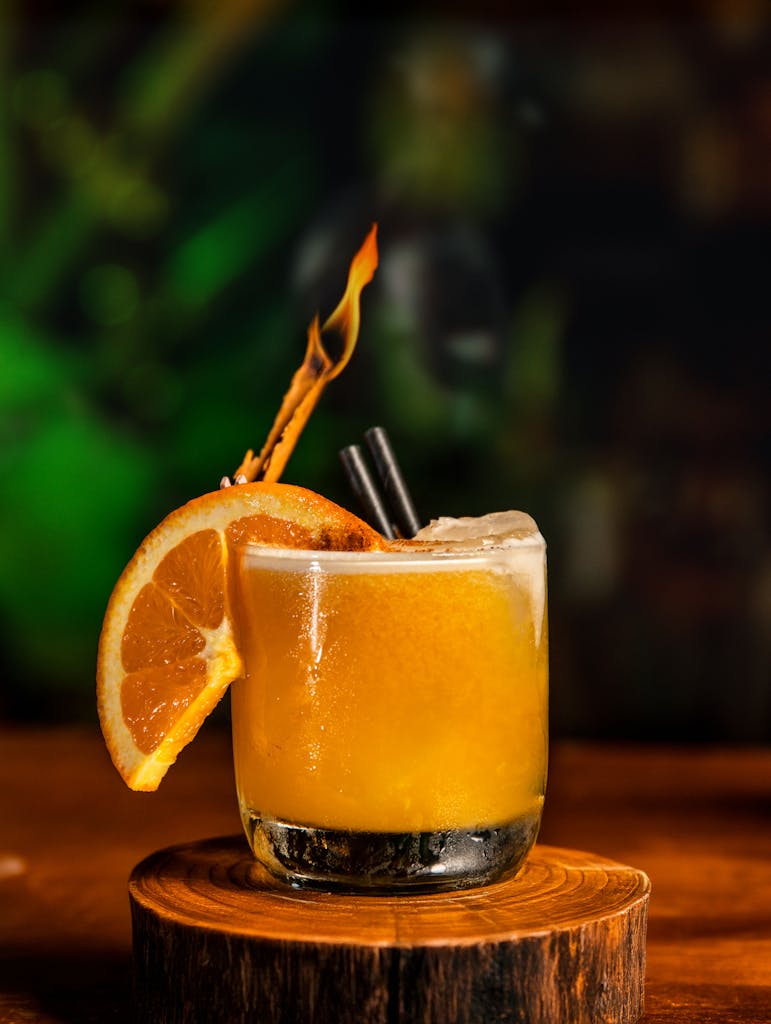 A refreshing orange cocktail with a flaming garnish on a rustic wooden coaster.