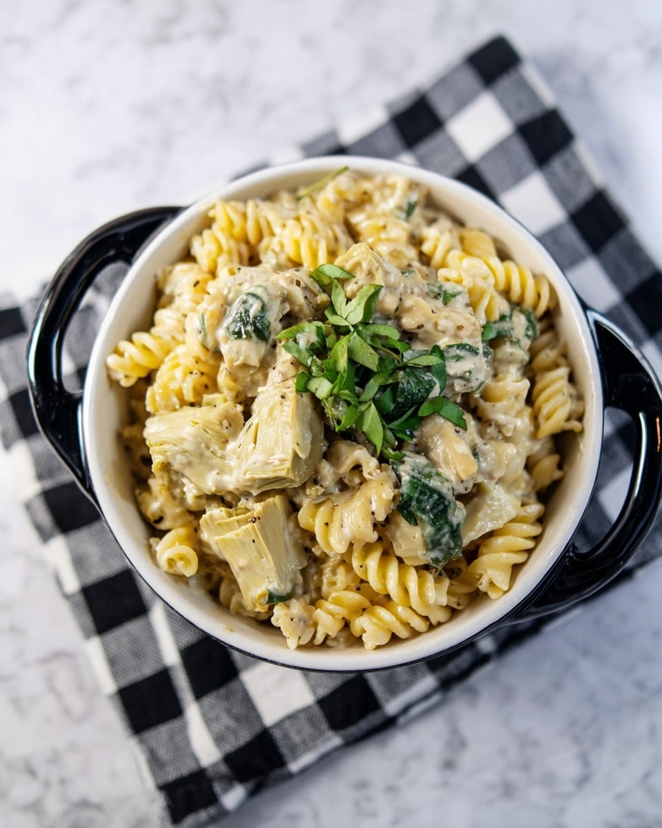 The image shows a bowl of creamy pasta with three visible layers: the bottom has light yellow spiral pasta, the middle is mixed with chunky artichoke pieces in pale green and creamy beige sauce, and the top is garnished with small, dark green spinach leaves. The white bowl with black handles is placed on a black and white checkered cloth, all set on a white marbled surface. The pasta looks soft and rich with a smooth, creamy texture, and the artichokes add a chunky element. photo taken with an iphone --ar 4:5 --v 7