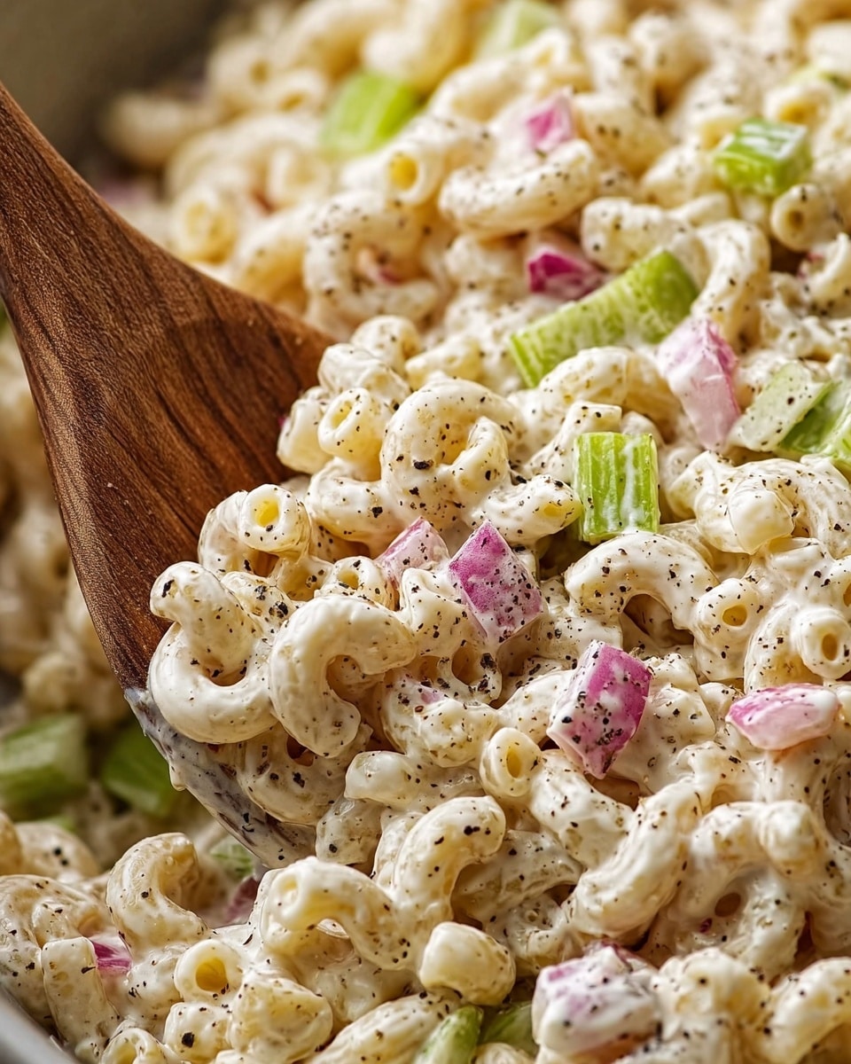 A close-up view of a creamy macaroni salad showing cavatappi pasta mixed with small pieces of red onion and crisp celery chunks evenly spread throughout. The salad is thickly coated in a white creamy dressing with a sprinkling of black pepper on top for texture and contrast. A wooden spoon is partially scooping some of the pasta salad, with its natural wood texture visible. The whole scene is set against a white marbled texture. photo taken with an iphone --ar 4:5 --v 7