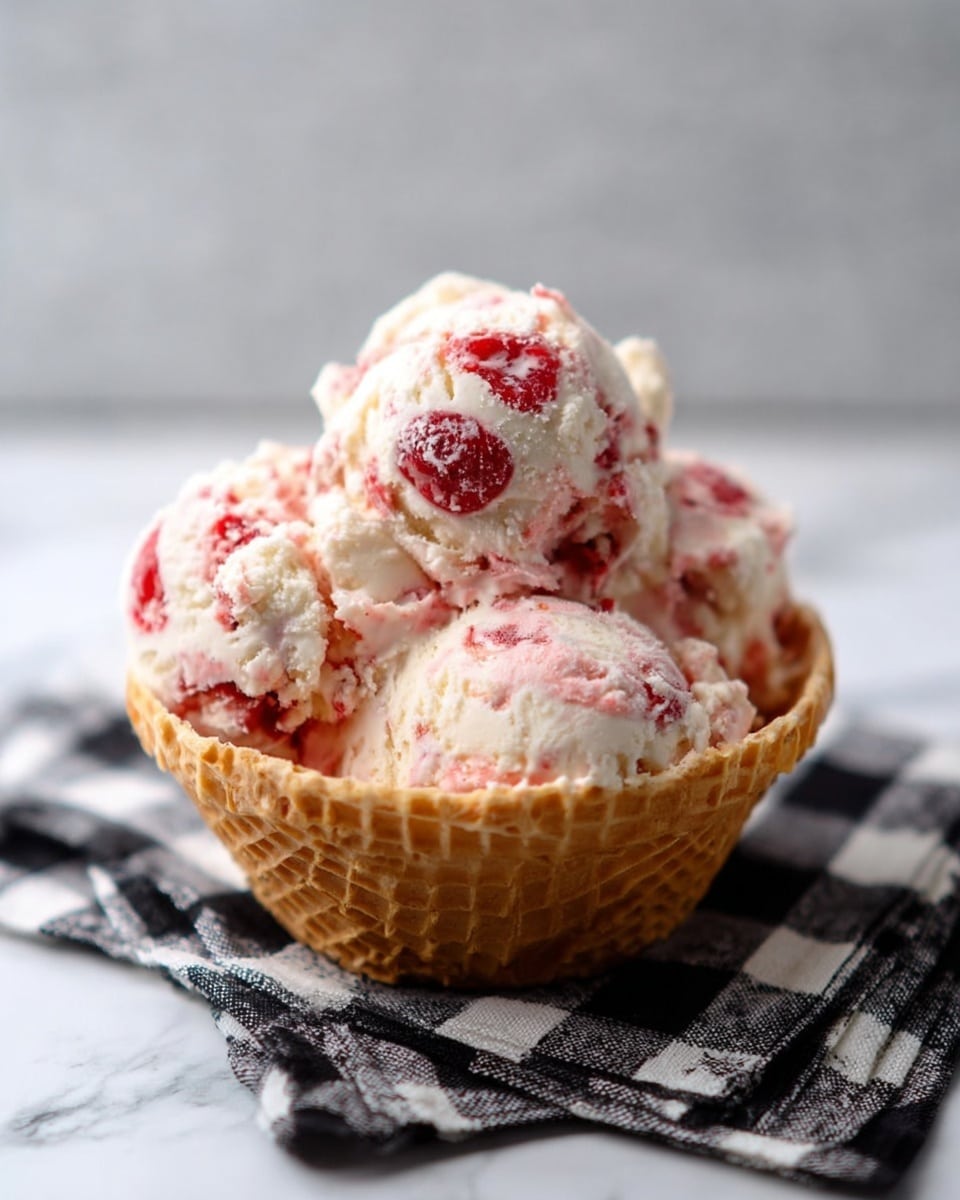 The image shows a waffle bowl filled with several scoops of creamy ice cream. The ice cream is white with pink swirls and has bright red fruit pieces mixed in. The texture looks soft with bits of the fruit visible throughout. The bowl sits on a black and white checkered cloth placed on a white marbled surface. The lighting is soft, highlighting the smooth and slightly melting texture of the ice cream. photo taken with an iphone --ar 4:5 --v 7
