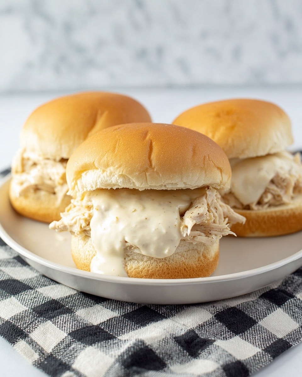Three sandwiches are placed on a white plate set on a black and white checkered cloth over a white marbled surface. Each sandwich has a soft, light brown bun with a smooth texture on top. Inside, there are shredded pieces of light beige chicken meat, topped with a generous amount of creamy white sauce that drips slightly from the sides. The chicken layer is thick and fills the lower half of the bun, while the sauce adds a moist texture. The three sandwiches are arranged close together, with the one in front being the most in focus. Photo taken with an iphone --ar 4:5 --v 7