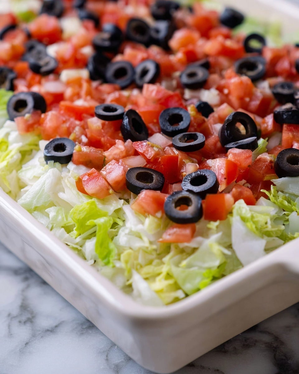 A close-up of a white square dish filled with a fresh salad consisting of three visible layers: the bottom layer is shredded light green lettuce with a soft, leafy texture; the middle layer is diced red tomatoes with a juicy appearance evenly spread over the lettuce; the top layer is sliced black olives arranged evenly, adding a dark, smooth texture. The dish sits on a white marbled surface. photo taken with an iphone --ar 4:5 --v 7