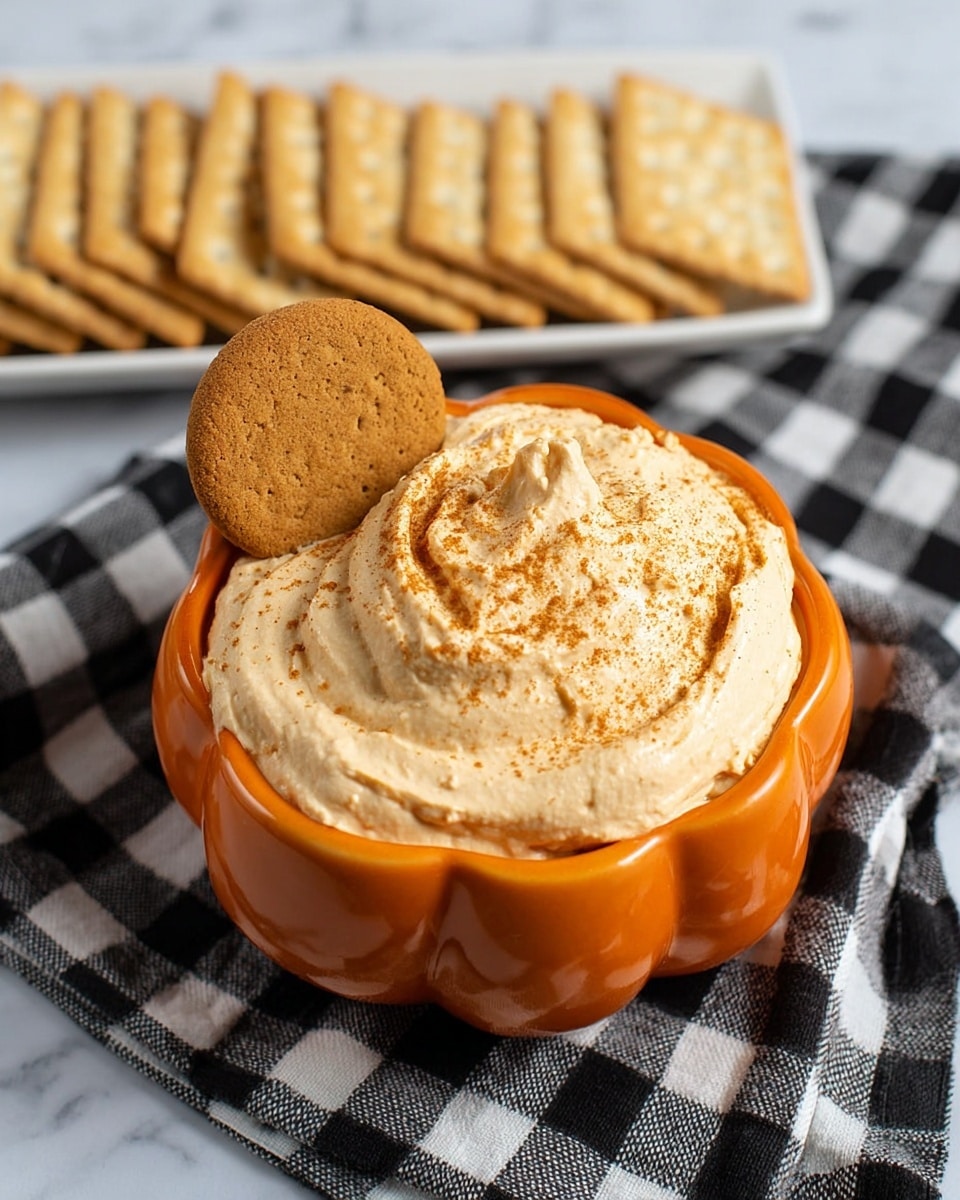 A small orange bowl shaped like a pumpkin holds a light orange creamy dip with a smooth, whipped texture and some brown powder sprinkled on top. One round cookie is placed standing inside the dip on the left side. In the background, there is a white rectangular plate filled with rectangular beige crackers. The bowl and plate sit on a black and white checkered cloth on a white marbled surface. Photo taken with an iphone --ar 4:5 --v 7