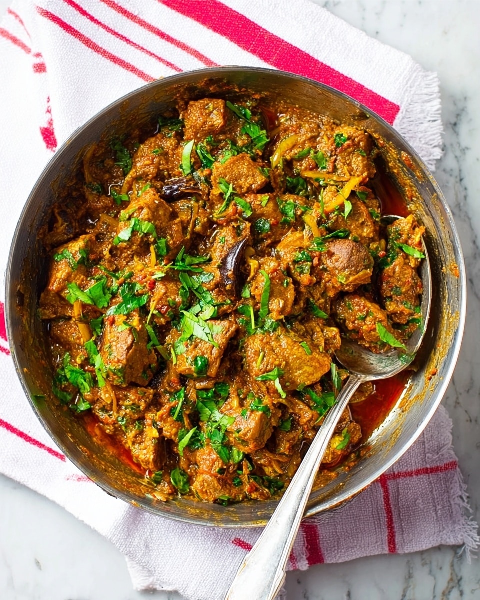 The image shows a round steel pan filled with a rich, chunky curry made of cooked meat pieces and green herbs. The meat is coated in a thick orange-brown sauce with visible onion slices and spices like a black pod. Bright green chopped cilantro is scattered on top, adding freshness and color contrast. The pan rests on a white cloth with red stripes, placed on a white marbled surface. A silver spoon with a long handle is resting inside the pan, partly buried in the food. photo taken with an iphone --ar 4:5 --v 7