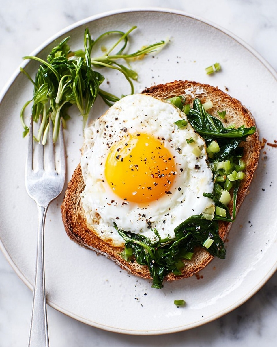 A white plate holds a single slice of toasted brown bread as the base layer, topped with a sunny-side-up fried egg with a bright yellow yolk and white edges. On the bottom right side of the egg, there are thin strips of cooked green leafy vegetables. Some extra green vegetable strips are placed beside the toast on the plate, and a silver fork rests nearby with a few vegetable pieces on its prongs. The plate sits on a white marbled surface with some black pepper sprinkled around. photo taken with an iphone --ar 4:5 --v 7