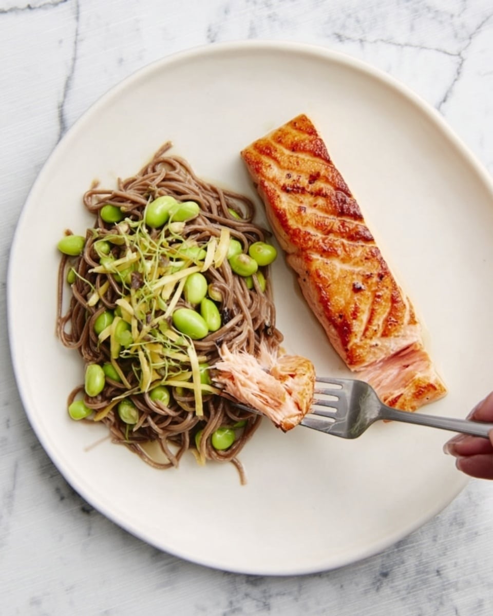 A white round plate holds two main parts: on the left, a small pile of brown soba noodles mixed with green edamame beans and light yellow baby corn, showing soft and fresh textures; on the right, a single piece of cooked salmon, golden brown on top with a slightly charred surface and a juicy inside where a piece has been cut. A silver fork is stuck into the pasta, lifting some noodles, and a woman's hand is holding the fork from the side. The plate rests on a white marbled surface, creating a clean and bright background. Photo taken with an iphone --ar 4:5 --v 7