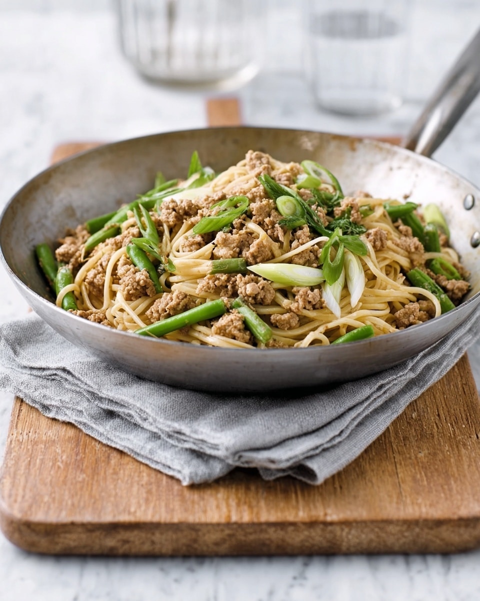A round silver pan filled with a noodle dish placed on a grey cloth napkin, which lies on a light brown wooden chopping board over a white marbled surface. The dish has a base layer of light brown noodles topped unevenly with cooked light brown minced chicken pieces and bright green sliced green beans, along with white and green chopped spring onions scattered on top. The pan has a sturdy metal handle extending out from the left side. Photo taken with an iphone --ar 4:5 --v 7