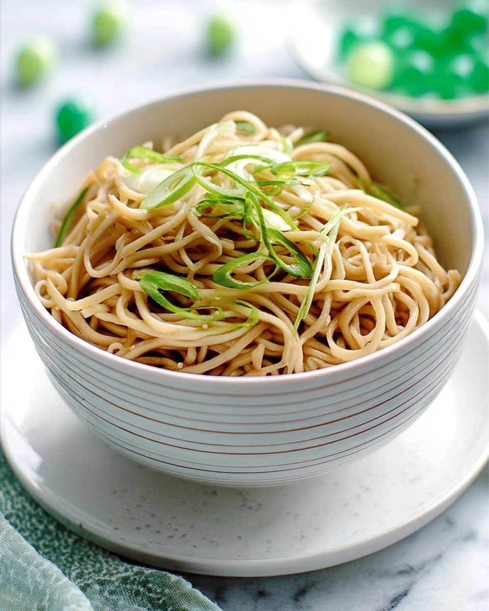 A white bowl filled with three layers of light brown noodles on top of each other, each layer showing a smooth and slightly glossy texture. Thin slices of green vegetables are mixed in and garnished on the top, adding a fresh vibrant color that contrasts with the noodles. The bowl is placed on a white marbled surface, with soft light shining from the side creating gentle shadows. Photo taken with an iphone --ar 4:5 --v 7