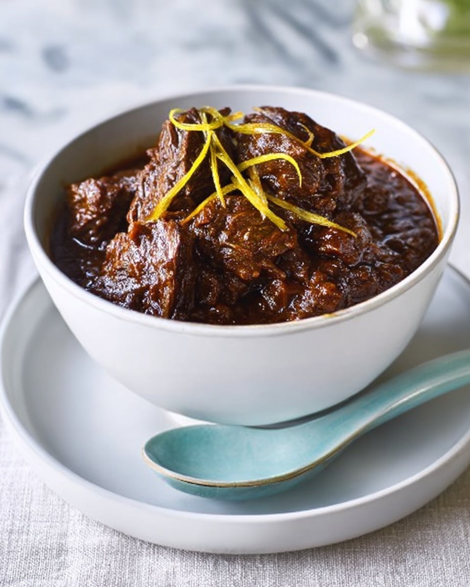 The image shows a white bowl filled with dark brown chunks of meat covered in thick, rich sauce with a slightly shiny texture. The meat looks tender and moist, with a few slivers of pale yellow garnish scattered on top. The bowl sits on a white plate, and next to it is a light blue ceramic spoon. The background is a white marbled surface. Photo taken with an iphone --ar 4:5 --v 7