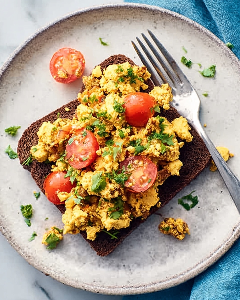 A piece of dark brown toast sits in the middle of a round white plate, topped with a colorful mix of soft scrambled yellow eggs mixed with bright red cherry tomato halves and specks of green herbs scattered on top. To the right of the toast, a silver fork rests partially on the plate. The plate is placed on a white marbled surface with a blue cloth partially visible. The colors are vibrant and fresh, showing a simple but tasty breakfast dish. photo taken with an iphone --ar 4:5 --v 7
