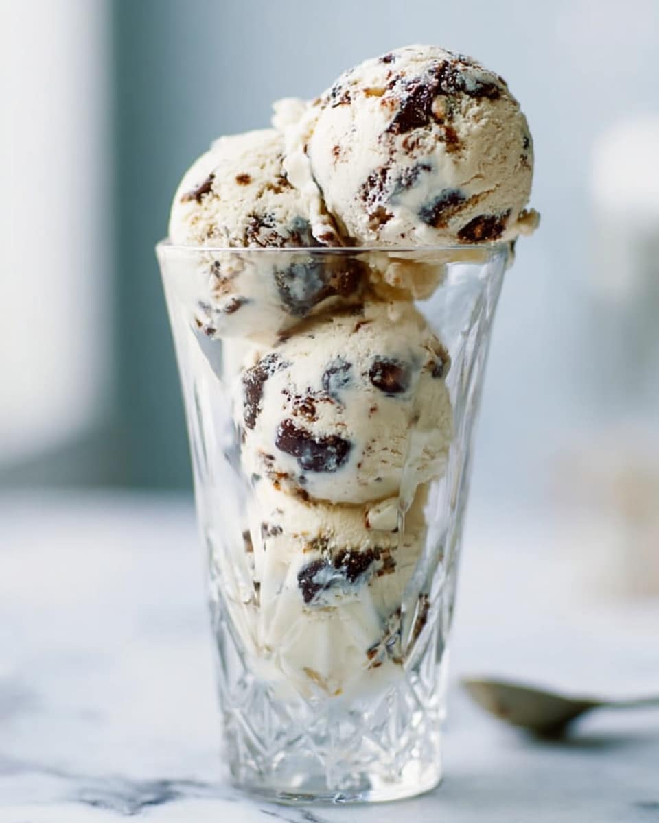 A clear glass cup filled with multiple scoops of light beige ice cream with visible dark brown and black chunks mixed throughout, forming a creamy textured dessert with a slightly uneven surface. The scoops are neatly stacked, creating about four layers from the bottom to the top of the cup. The background shows a soft focus on cool white and light tones, set on a white marbled surface with some kitchen utensils blurred in the back. photo taken with an iphone --ar 4:5 --v 7