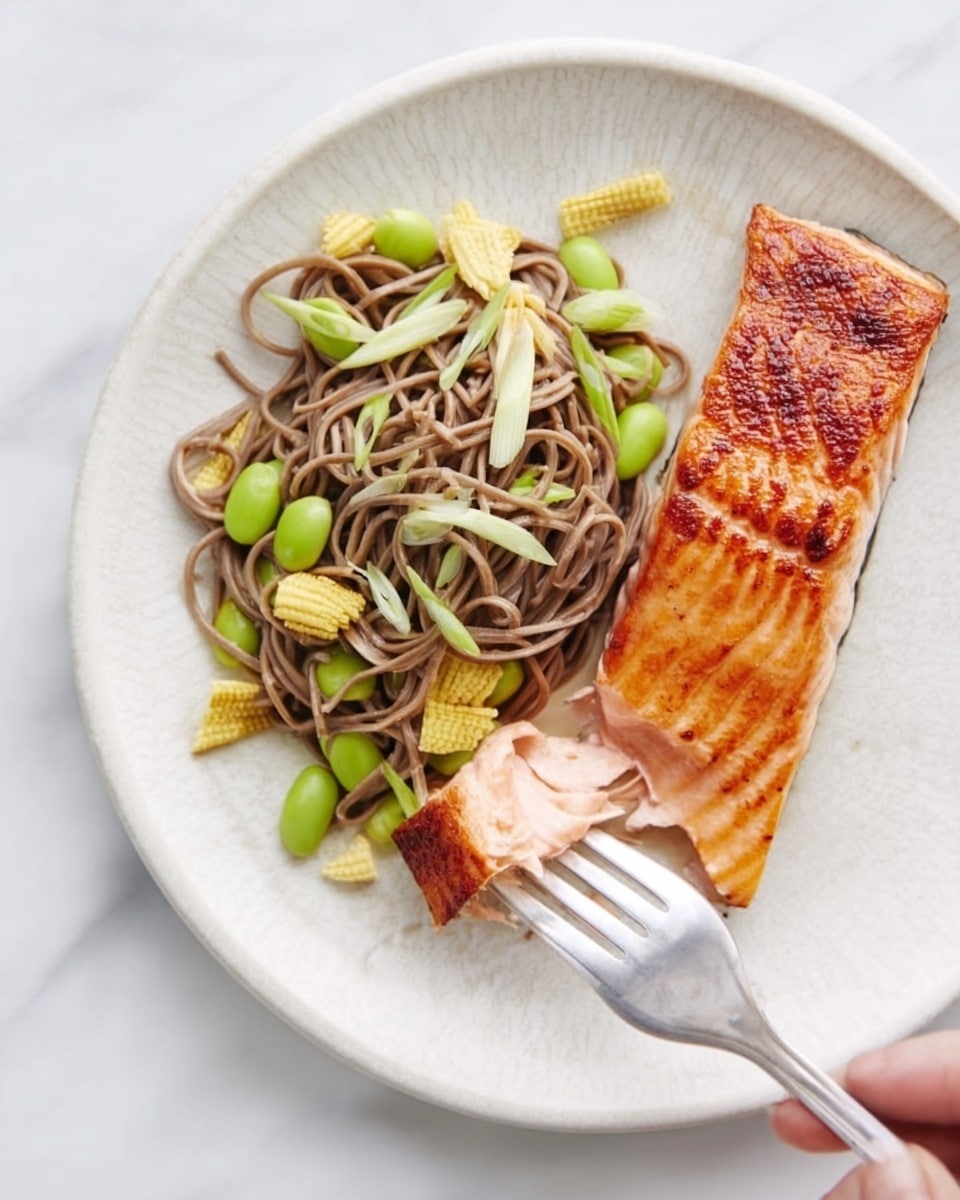 The image shows a white plate with two main parts: on the left, a pile of soba noodles mixed with green vegetables like edamame and baby corn, creating a mix of light brown and green colors with a soft texture; to the right, a piece of cooked salmon with a golden-brown, glossy surface and firm texture; a fork held by a woman's hand is lifting a small piece of salmon from the right side of the plate; the background is a white marbled texture. photo taken with an iphone --ar 4:5 --v 7