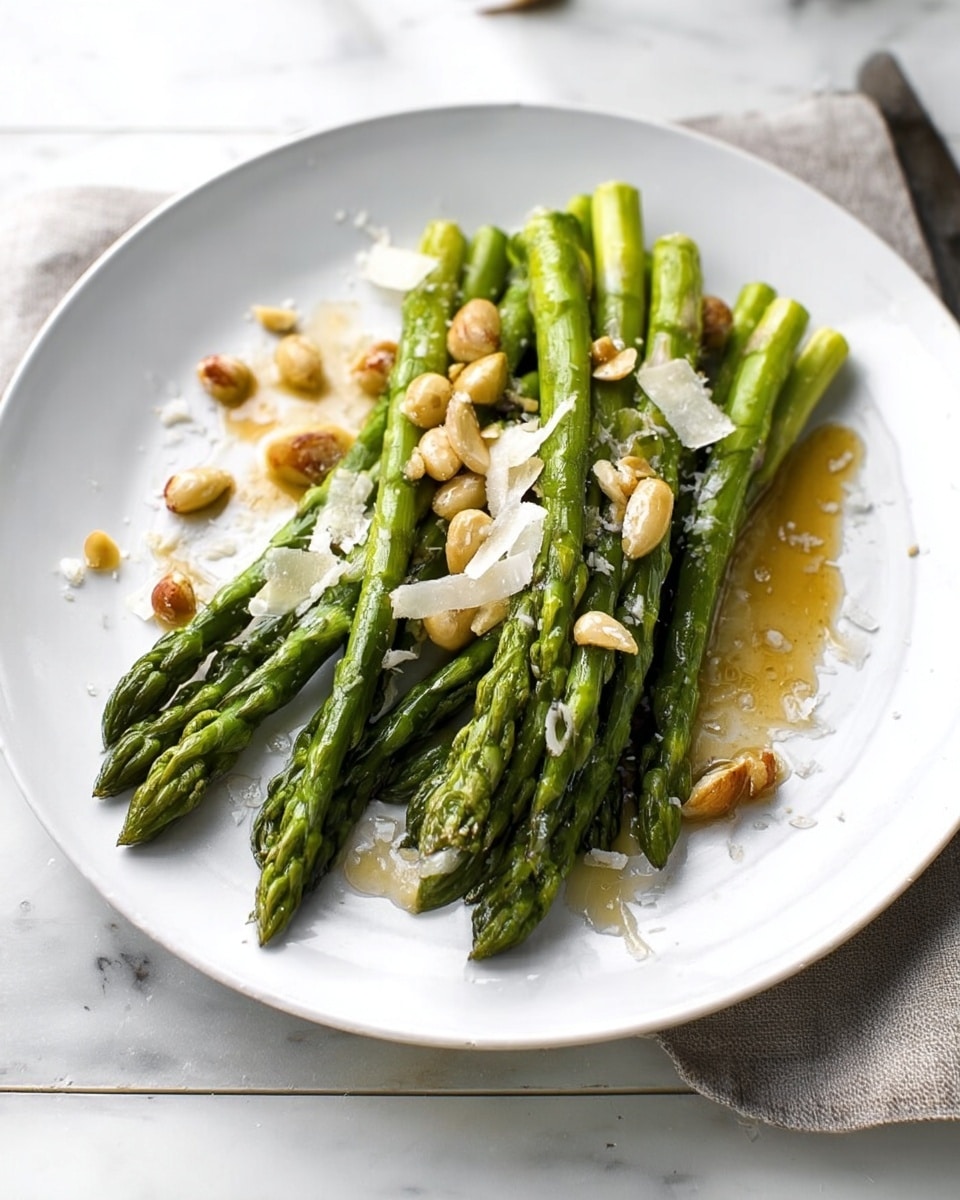 A white plate holds a small pile of bright green asparagus spears arranged in a neat stack with tips pointing mostly to the right. Around and slightly under the asparagus are light brown toasted nuts and small white shavings that look like cheese. The dish is lightly drizzled with a shiny golden sauce that pools a little on the plate. The setting includes a wooden table beneath the plate and a blurry background with a lemon wedge and cutlery visible. The overall look is fresh and simple with natural colors. photo taken with an iphone --ar 4:5 --v 7