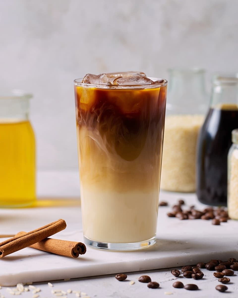 A clear glass filled with a three-layer iced coffee drink stands on a white marbled surface. The bottom layer is creamy white with a thick texture, the middle is a light beige with smooth blending, and the top layer is dark brown coffee with ice cubes floating and light milk dripping down the side. Next to the glass are two cinnamon sticks lying crossed, scattered coffee beans, white rice grains, and two small clear glass containers, one with dark coffee and the other with pale yellow cream. The background is a soft light bluish-white with subtle texture. Photo taken with an iphone --ar 4:5 --v 7