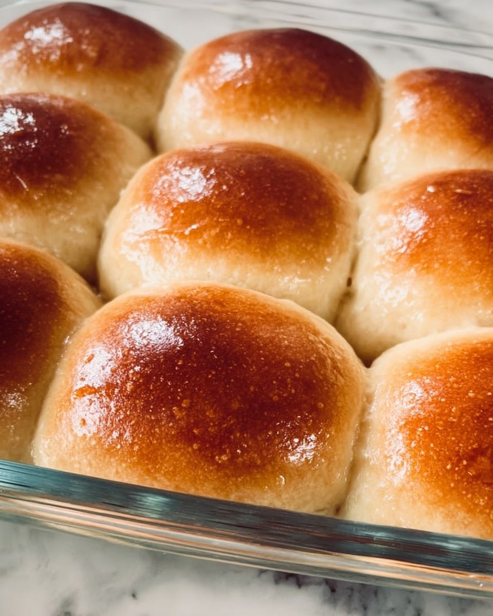 The image shows a close-up of a glass baking dish filled with freshly baked dinner rolls arranged in three rows with soft, light golden-brown tops that are slightly shiny, and pale sides pressed closely together. The rolls have a smooth texture with a slight gradient of color from darker brown on the crown to light beige on the sides. The dish sits on a white marbled surface, adding a clean and bright contrast to the warm tones of the bread. Photo taken with an iphone --ar 4:5 --v 7