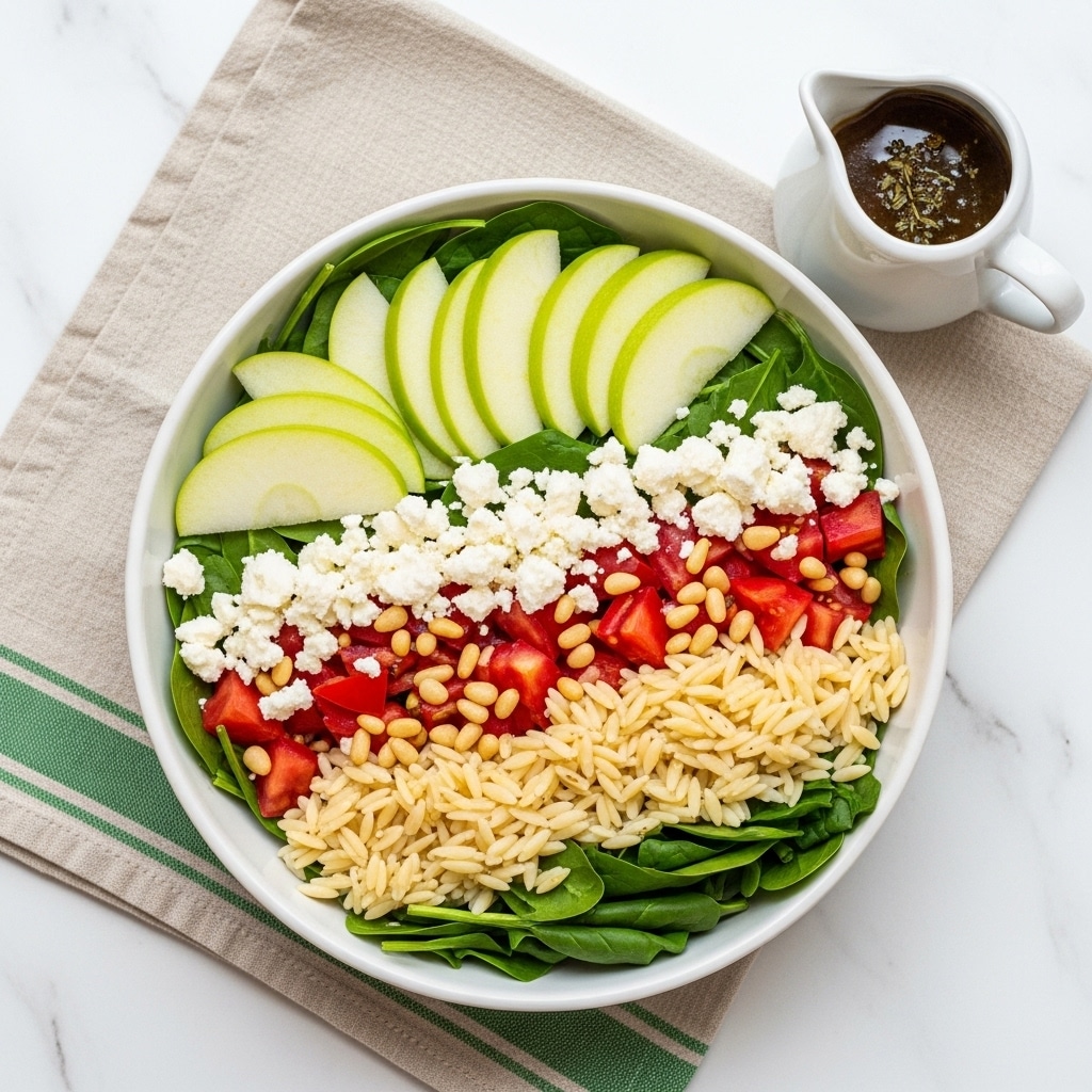 A white bowl filled with a fresh salad sits on a beige cloth with green stripes, placed on a white marbled texture surface. The salad has multiple layers visible: the bottom layer has small brown orzo pasta, mixed with chopped dark green leafy spinach. Scattered throughout are bright red diced tomatoes, light green thin apple slices, and small crumbles of white feta cheese. Light brown pine nuts are also sprinkled evenly on top. To the upper left of the bowl is a small white sauce container filled with dark-colored dressing that looks like a mix of oil and herbs. The photo taken with an iphone --ar 4:5 --v 7
