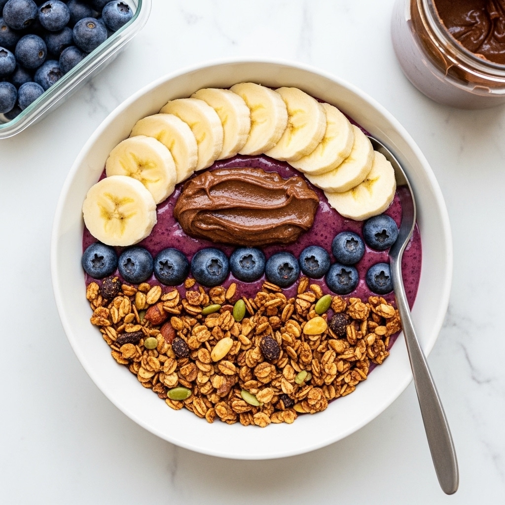 A white bowl holds a layered dish starting with a base of granola covering half of the bowl, showing a rough, crunchy texture in golden brown shades. On top of the granola sits a small cluster of dark blue, smooth blueberries near the center. Surrounding the blueberries is a small dollop of thick, dark brown spread with a rich, creamy texture. Arranged neatly along the opposite half of the bowl are six slices of pale yellow banana with a smooth and slightly creamy look. The bowl rests on a white marbled surface, with part of a silver spoon placed on the edge of the bowl. A clear container with more dark blueberries is seen top left and part of a small jar with dark brown contents on the top right. photo taken with an iphone --ar 4:5 --v 7