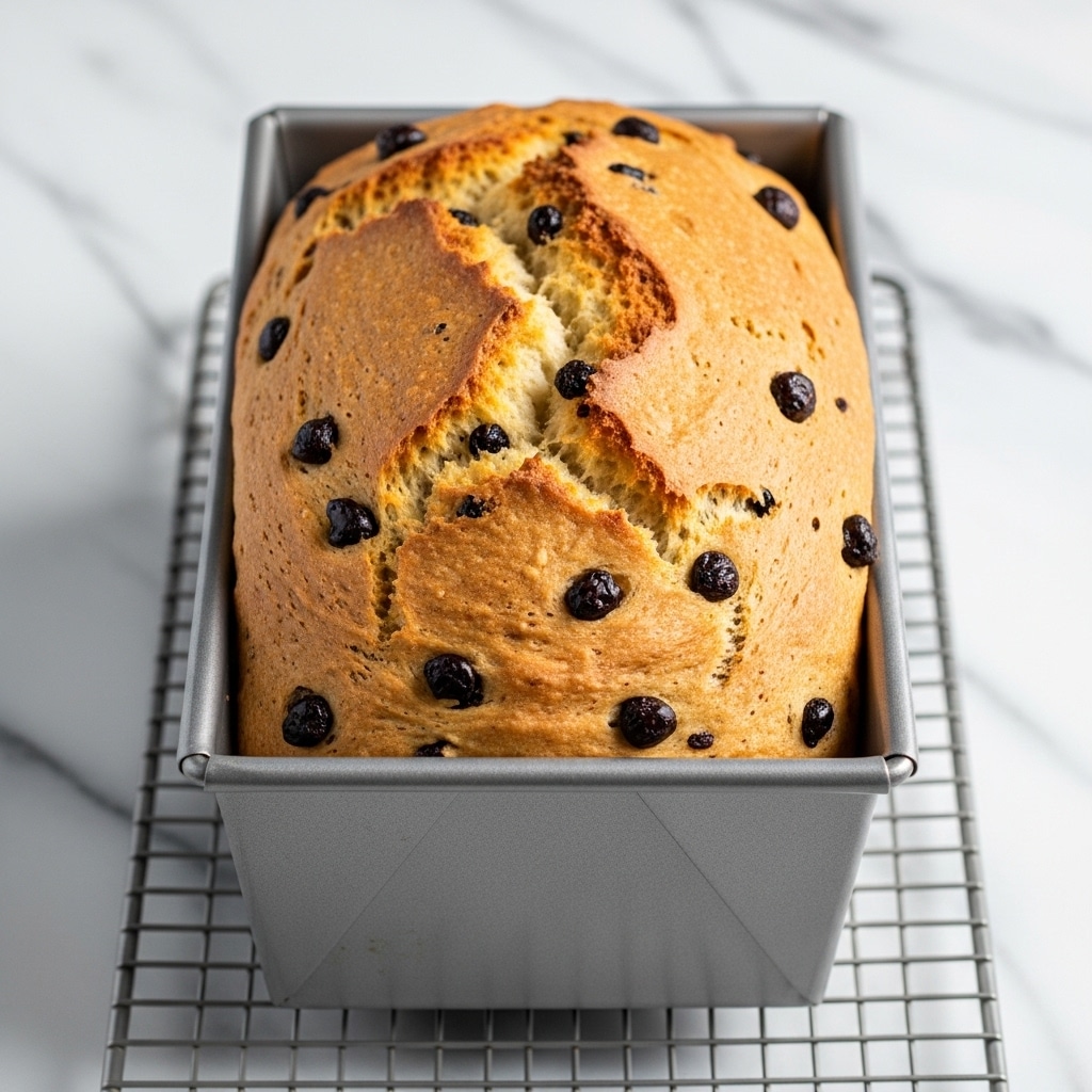 The image shows a freshly baked loaf of bread in a silver metal baking pan resting on a rack inside an oven. The bread has a golden brown crust with a few cracks on the top, revealing a soft inside with dark spots that look like raisins or chocolate chips scattered throughout. The texture of the crust looks firm and slightly rough, while the inside appears moist and dense. The background is a white marbled texture that contrasts gently with the warm tones of the bread. Photo taken with an iphone --ar 4:5 --v 7