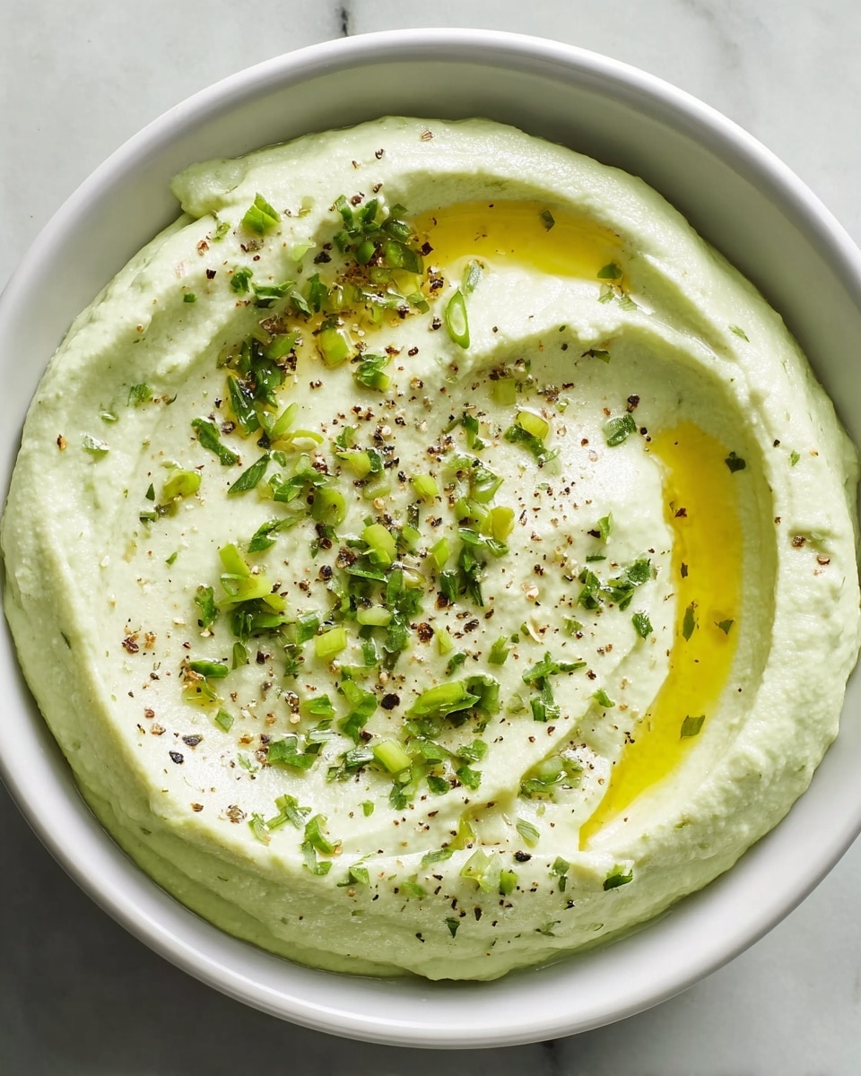 A white bowl filled with one thick layer of pale green creamy dip, swirled with a smooth texture. On top, there is a drizzle of golden yellow olive oil, thinly spread in a curve. Scattered over the dip are small pieces of finely chopped green herbs and a light sprinkle of coarse black pepper and salt. The bowl sits on a white marbled surface. photo taken with an iphone --ar 4:5 --v 7
