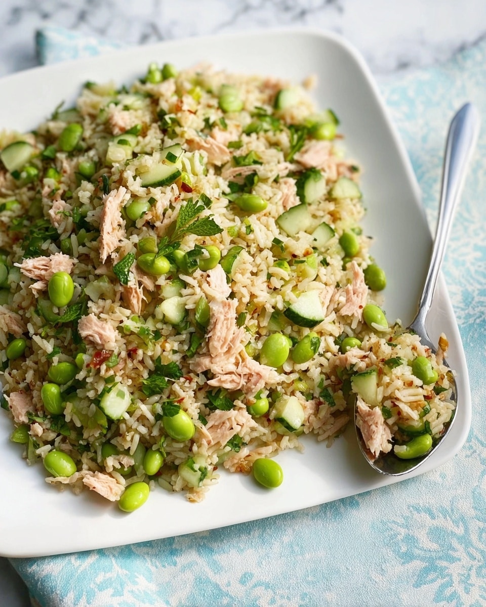 A white plate is filled with a colorful rice salad layered with light brown cooked rice, bright green edamame beans, sliced cucumbers, chopped leafy greens, and small pieces of pinkish cooked chicken, all mixed together. On the right side of the plate, a silver spoon rests on the edge with some of the salad on it. The dish is placed on a white marbled surface with a light blue and white patterned cloth partially visible underneath. Photo taken with an iphone --ar 4:5 --v 7