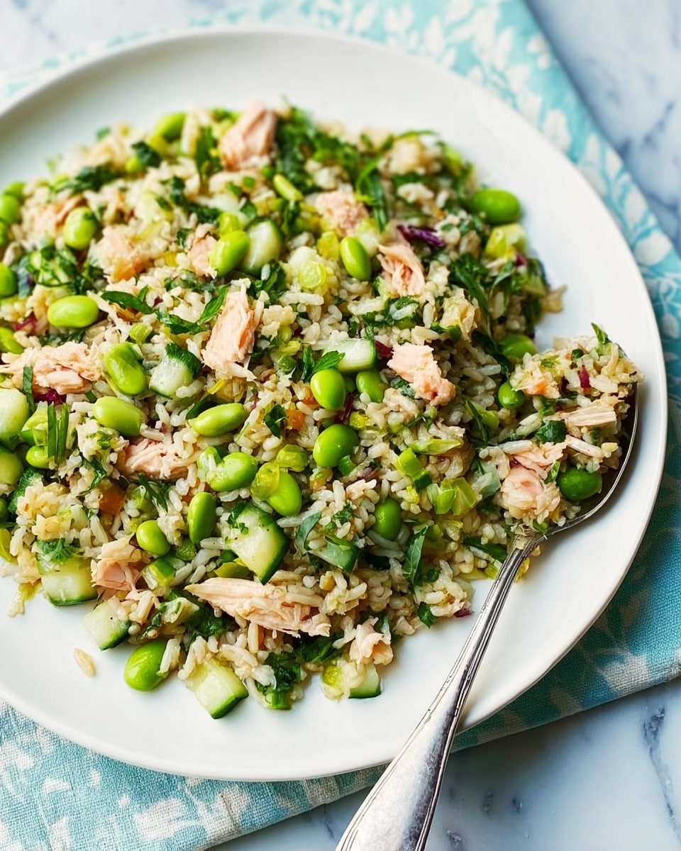 A white square plate filled with a mixed rice salad that has three main layers: a base layer of light brown rice grains, scattered green edamame beans and chopped cucumber pieces making up the middle, and pieces of light pink tuna mixed evenly throughout. The salad looks fresh with some small bits of green herbs visible, adding texture and color. On the right side of the plate, a silver spoon holds a small scoop of the salad, resting partly on the plate. The plate is placed on a white marbled surface with a soft blue and white patterned cloth partially underneath. Photo taken with an iphone --ar 4:5 --v 7