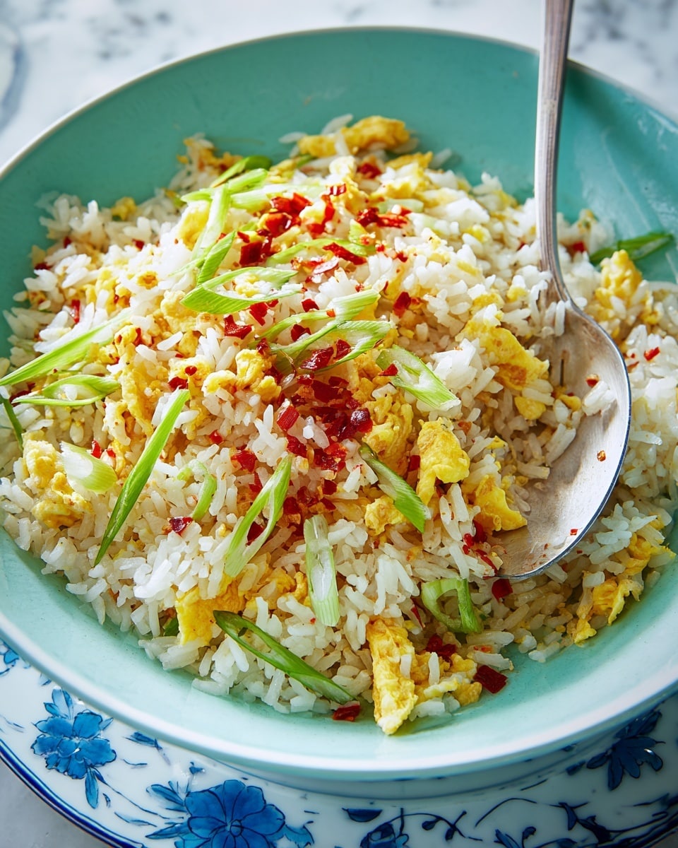 A close-up of a white bowl with a blue inside filled with fried rice. The fried rice has about three main layers: the white rice grains mixed with small yellow scrambled egg pieces scattered throughout, thinly sliced green onions layered on top and mixed in, and tiny bright red chili flakes sprinkled across the dish. A silver spoon rests inside the bowl holding some rice and onions, and the bowl sits on a white plate with a few pieces of green onion around it, all placed on a white marbled surface. Photo taken with an iphone --ar 4:5 --v 7