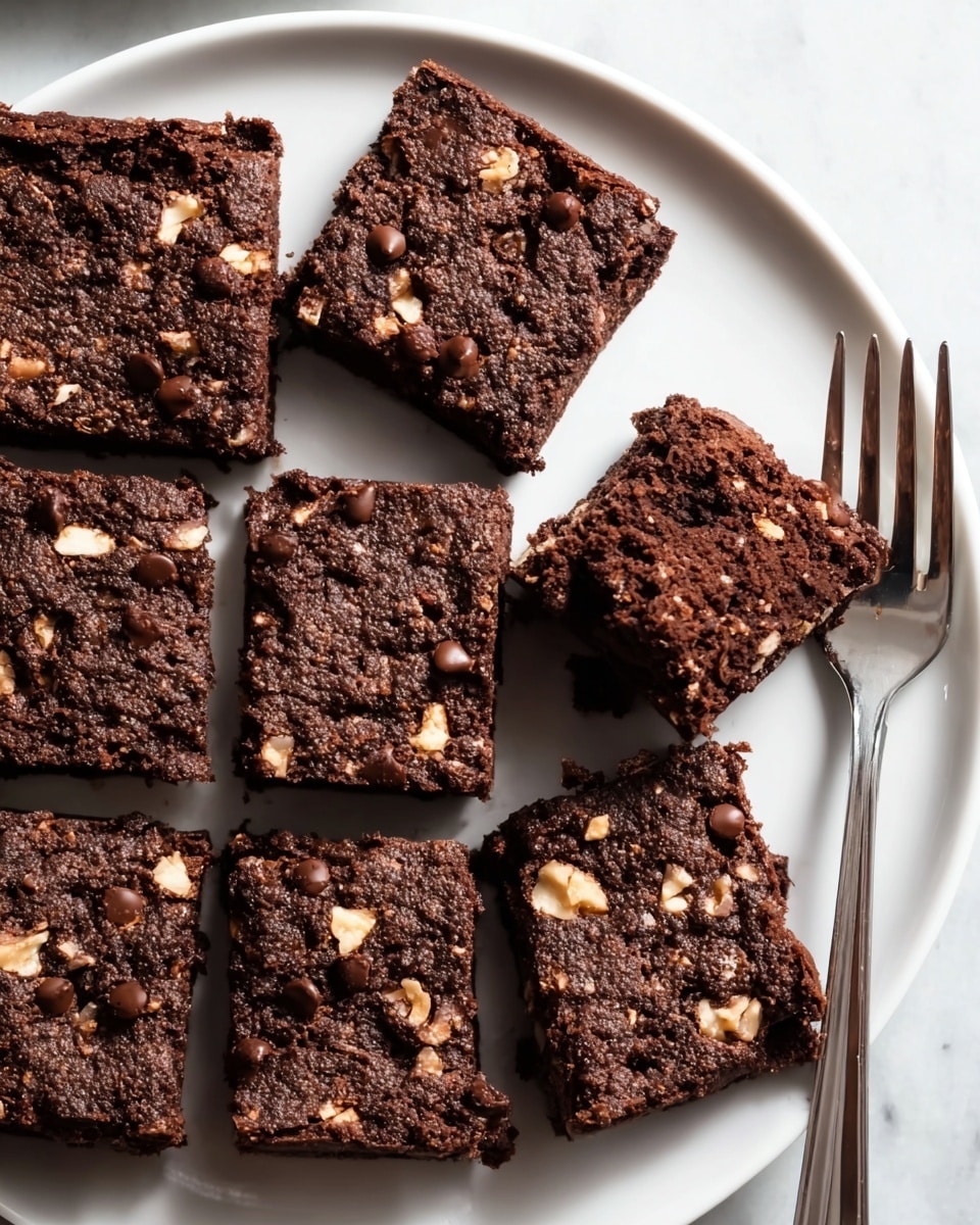 The image shows nine square pieces of chocolate brownies arranged closely on a large white plate with a shiny metal fork placed beside them. Each brownie is thick and dense with a dark brown color, speckled generously with small black chocolate chips and chunks of light tan walnuts embedded throughout the top and inside texture, giving a rough and slightly crumbly surface. The brownies appear moist, firm, and rich, with clean even edges, sitting on a smooth white marbled texture background. Photo taken with an iphone --ar 4:5 --v 7