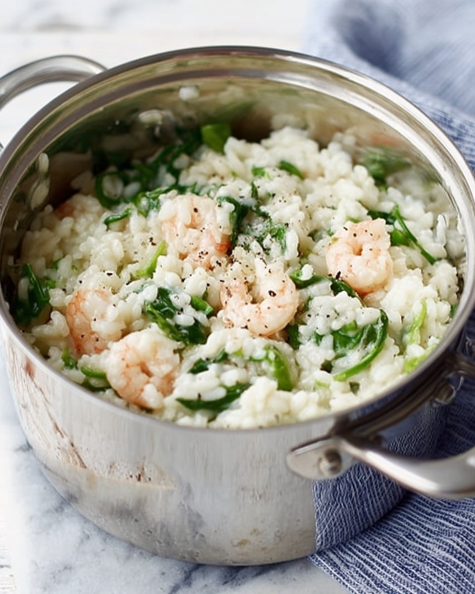 The image shows a close-up of a shiny silver pot filled with risotto. The risotto has creamy white rice with a soft, slightly sticky texture mixed with pieces of green leafy vegetables and small cooked shrimp that are light pink with a slightly translucent look. The top is sprinkled with small freshly ground black pepper spots. The pot is set on a white marbled surface, and a soft, folded blue and white cloth is placed in the background. Photo taken with an iphone --ar 4:5 --v 7