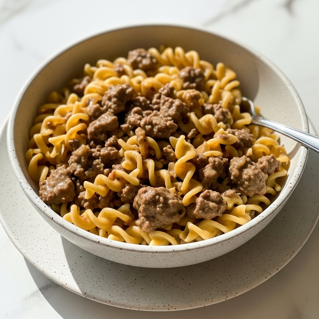 A close-up view of a bowl filled with creamy beef stroganoff made from egg noodles and ground beef in a light brown sauce. The noodles are soft and twisted, mixed evenly with small chunks of browned beef, all coated in a smooth, slightly glossy sauce. The dish is served in a speckled white bowl resting on a matching speckled white plate. There is a silver fork placed on the right side inside the bowl. The background is a white marbled texture with some soft natural light creating a warm tone. photo taken with an iphone --ar 4:5 --v 7