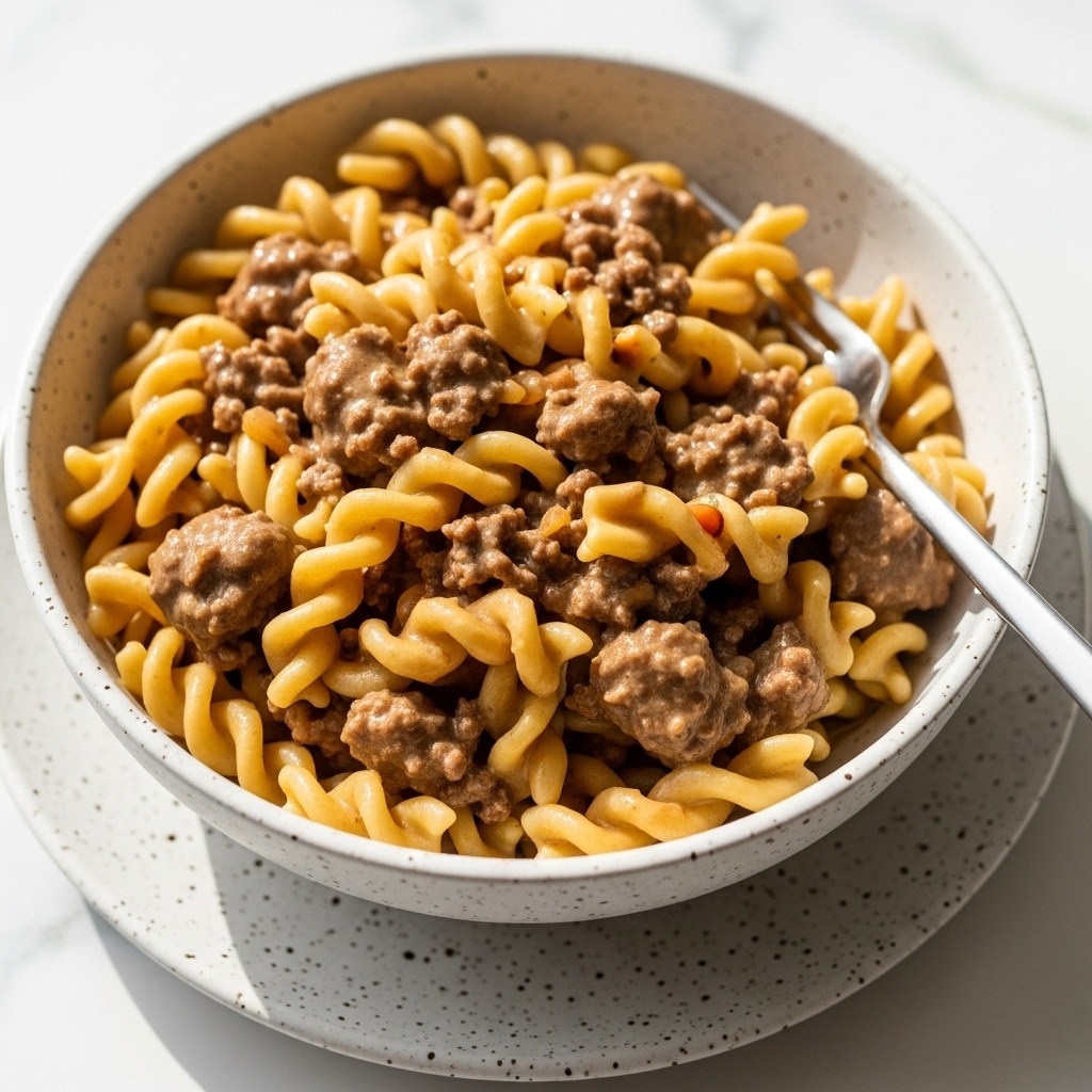 A close-up of a bowl filled with creamy beef stroganoff made of pasta noodles and ground beef in a brown sauce. The pasta has a pale yellow color with visible spirals mixed evenly with small crumbled pieces of brown beef. The sauce covers the pasta and beef, giving a glossy and slightly thick texture. The round bowl is white with small brown speckles, resting on a white marbled surface, with a fork placed inside the bowl on the right side. photo taken with an iphone --ar 4:5 --v 7