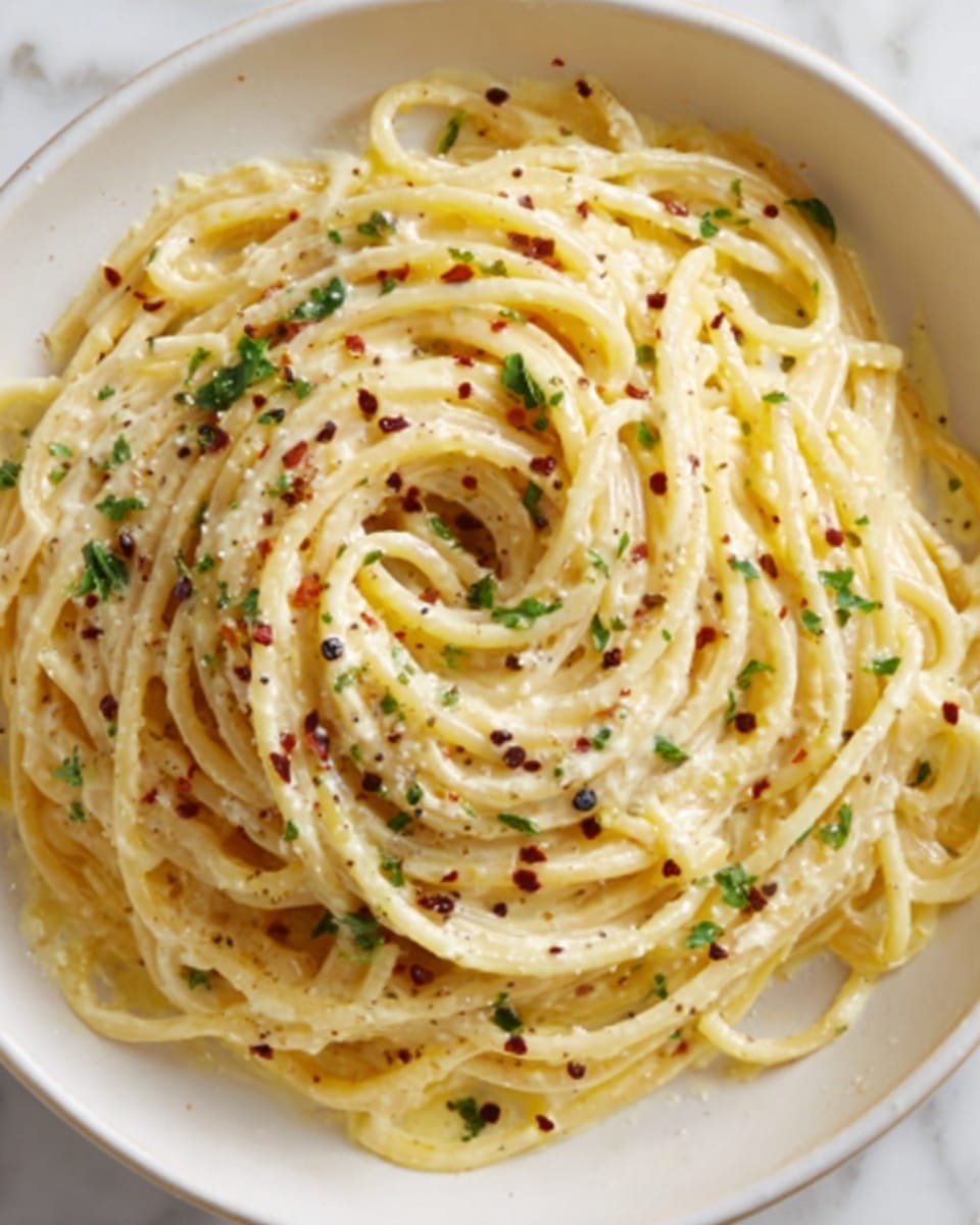 A white bowl filled with spaghetti noodles coated in a creamy yellow sauce, with small bits of green herbs and red pepper flakes scattered evenly over the top. The noodles are loosely twirled in layers, showing a smooth and slightly glossy texture, with some strands overlapping each other softly. The background is a white marbled surface. Photo taken with an iphone --ar 4:5 --v 7