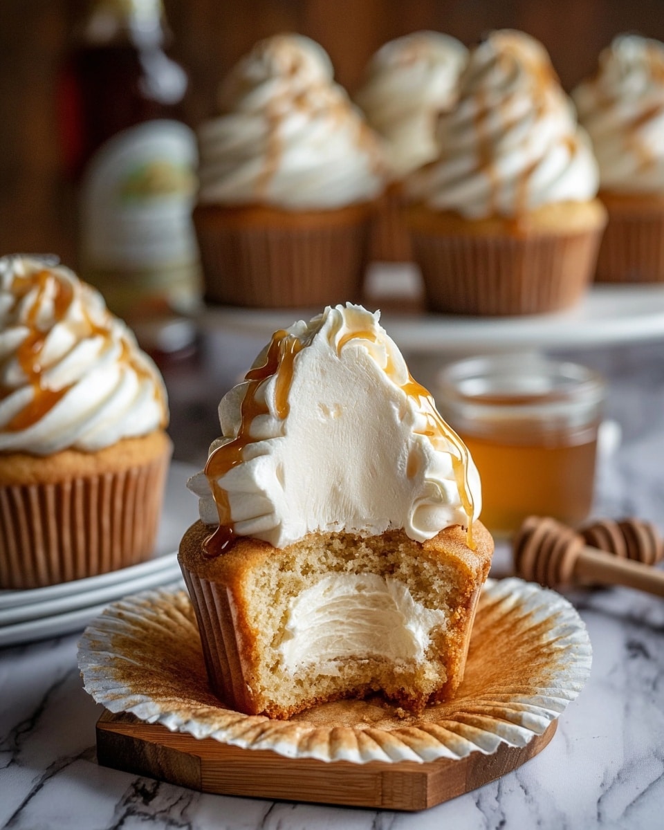 The image shows a close-up of a cupcake with two main layers. The bottom layer is a golden-brown cupcake with a soft, crumbly texture visible inside, as it has a big bite taken out of it. On top, there is a tall swirl of creamy white frosting with a smooth, fluffy texture and light caramel drip on it. The cupcake sits on an unwrapped white paper liner, placed on a wooden board. In the background, there are more cupcakes with the same frosting and caramel drizzle, all sitting on a white plate, with a white marbled surface underneath. Photo taken with an iphone --ar 4:5 --v 7