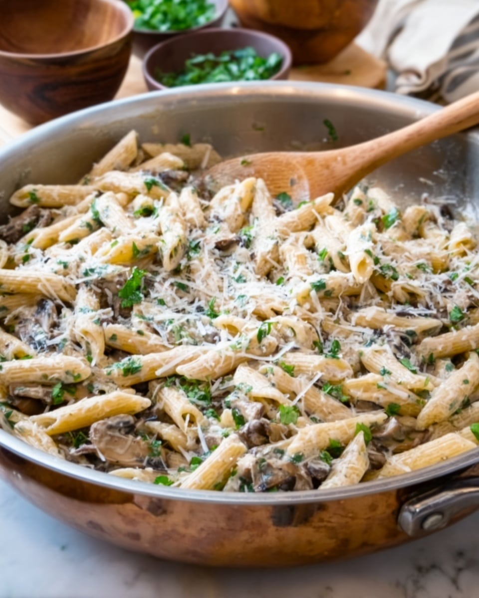 A large silver pan filled with penne pasta coated in a creamy white sauce, mixed with small pieces of cooked mushrooms and sprinkled with finely chopped green herbs and grated cheese on top. The pasta has a light beige color with bits of dark brown mushrooms spread evenly throughout. The green herbs add fresh spots of color, and the cheese gives a slightly grainy texture on the surface. The pan sits on a white marbled surface with some blurred ingredients in the background. Photo taken with an iphone --ar 4:5 --v 7