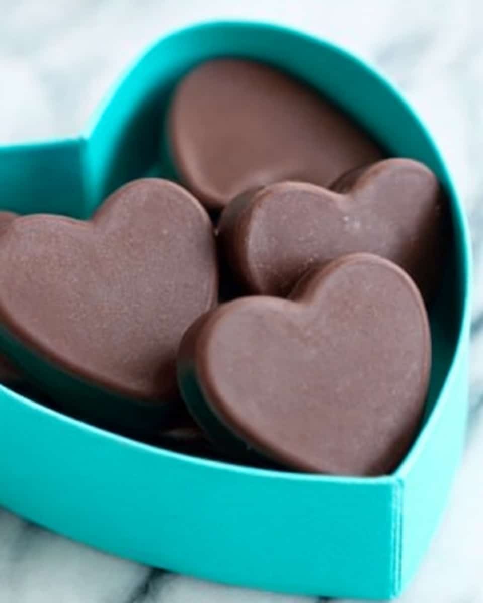 The image shows a small white box filled with several heart-shaped chocolates. Each chocolate is smooth with a matte dark brown color and neat edges. The chocolates are tightly packed inside the box, slightly stacked so some are leaning on others. The box has a bright turquoise lid partially visible, creating a bright contrast with the dark chocolates. The background has a white marbled texture. photo taken with an iphone --ar 4:5 --v 7