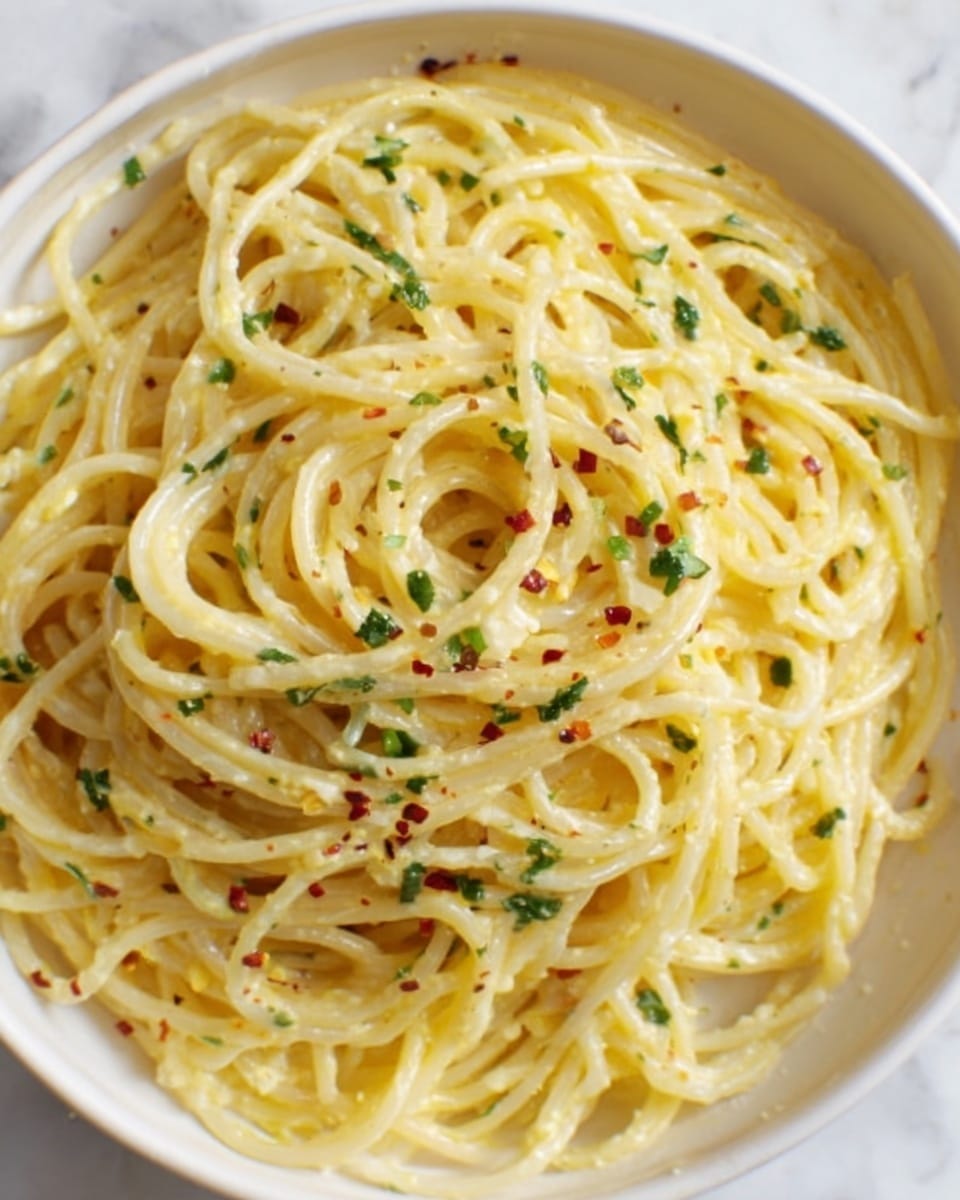A white bowl filled with a single layer of creamy spaghetti piled neatly in the center, coated in a pale yellow sauce with visible specks of black pepper and red chili flakes scattered evenly on top. Fresh green herbs are lightly sprinkled throughout, adding small pops of color against the smooth sauce. The texture looks slightly glossy and rich, with a few strands of pasta slightly curled and overlapping. The bowl sits on a white marbled surface. photo taken with an iphone --ar 4:5 --v 7