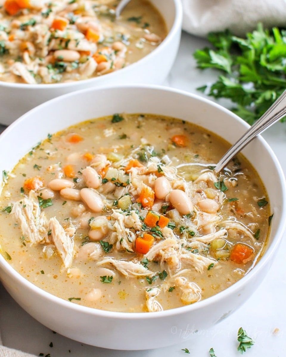 A white bowl filled with thick chicken and bean soup sits on a white marbled surface. The soup has a beige, creamy broth with visible chunks of shredded white chicken, small orange carrot pieces, pale beans, and bits of green parsley scattered on top. A silver spoon rests inside the bowl on the left side. In the background, another white bowl with the same soup is slightly out of focus with some green parsley visible behind it. photo taken with an iphone --ar 4:5 --v 7