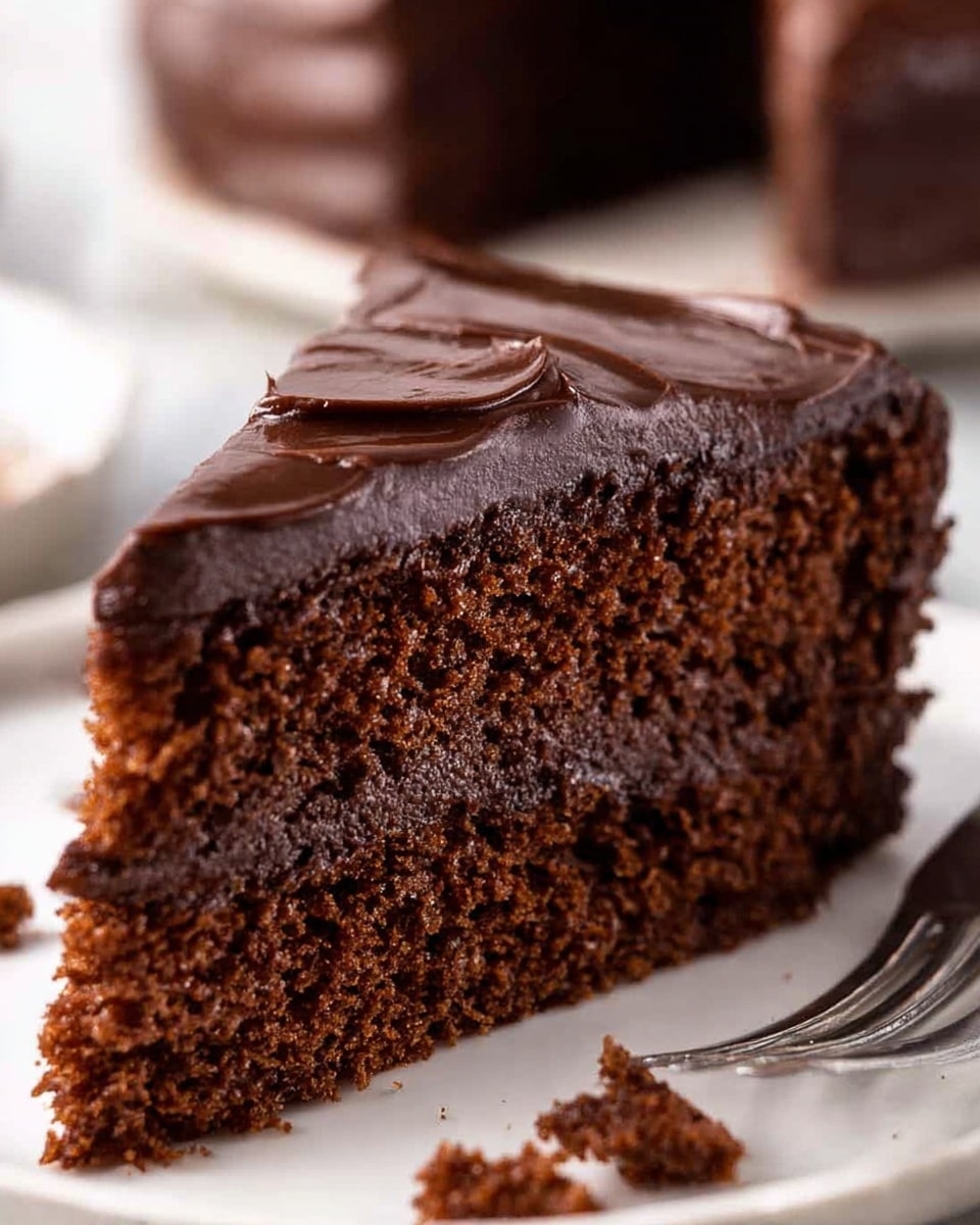 A close-up image of a single slice of chocolate cake with two layers: the bottom layer is a moist, rich brown cake with a soft, crumbly texture, and the top layer is a thick, dark chocolate frosting with smooth swirls and waves covering the entire top surface. The slice is placed on a white plate on a white marbled textured surface, with chocolate crumbs scattered around it and a silver fork blurred in the background photo taken with an iphone --ar 4:5 --v 7