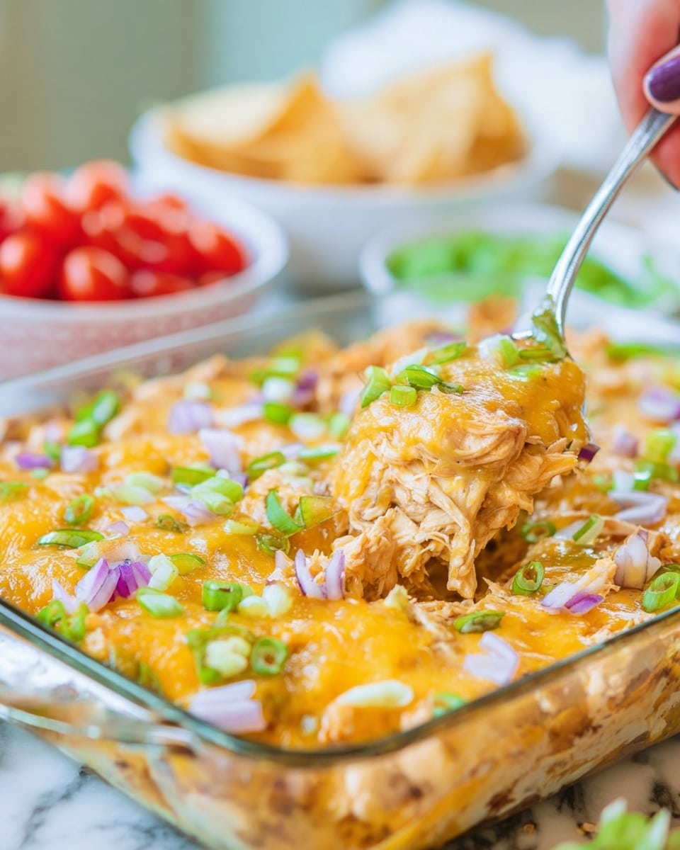 A clear glass dish filled with a layered cheesy chicken casserole; the bottom layer consists of shredded chicken pieces mixed in a creamy sauce, topped with a thick melted layer of golden cheddar cheese that covers the dish unevenly, sprinkled with chopped green onions and thin slices of red onion, giving pops of green and purple on top. A silver spoon is lifting a portion from one corner, showing the cheesy, moist, and textured layers. The dish sits on a white marbled surface with blurred bowls of cherry tomatoes and green leaves in the background. Photo taken with an iphone --ar 4:5 --v 7
