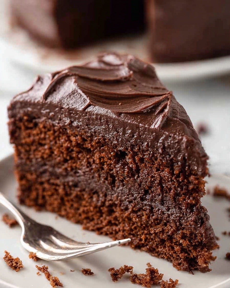 A close-up view of a single slice of chocolate cake on a white plate, showing two layers: the bottom layer is a light, fluffy dark brown chocolate cake with a moist texture, topped by a thick, smooth layer of glossy dark chocolate frosting with subtle swirls on top. In front of the slice, some crumbs and a small piece of cake lie scattered, and a fork rests blurred in the background on a white marbled surface. photo taken with an iphone --ar 4:5 --v 7