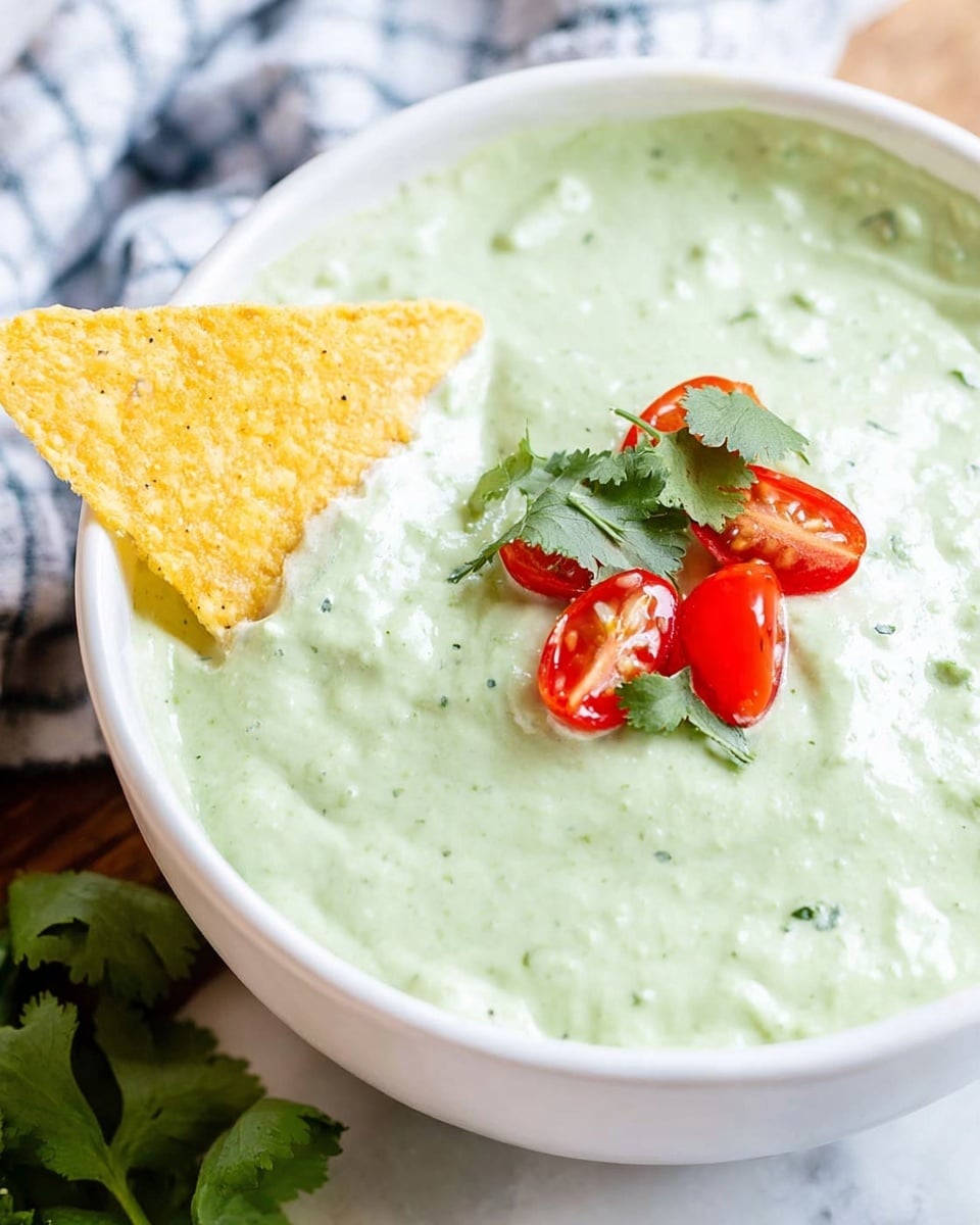 A white bowl filled with smooth, creamy light green sauce with tiny dark specks, topped with small bright red tomato pieces and fresh green cilantro leaves placed in the center. A yellow tortilla chip dipped into the sauce is held by a woman's hand, showing the thick, creamy texture of the sauce dripping from the chip. The bowl is set on a white marbled surface with soft natural light brightening the scene. photo taken with an iphone --ar 4:5 --v 7