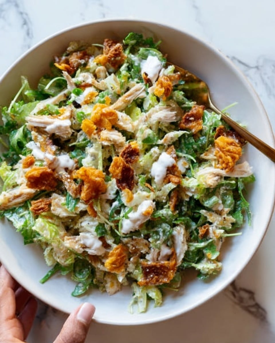 A white plate filled with a colorful salad layered in a casual mix; the base is green with chopped leafy vegetables and arugula, topped with shredded white chicken pieces scattered unevenly. There are golden-brown crispy fried bits spread around, adding a crunchy texture, and dollops of creamy white dressing placed in small spots on top. A woman's hand is gently holding the edge of the plate over a white marbled surface. Photo taken with an iphone --ar 4:5 --v 7