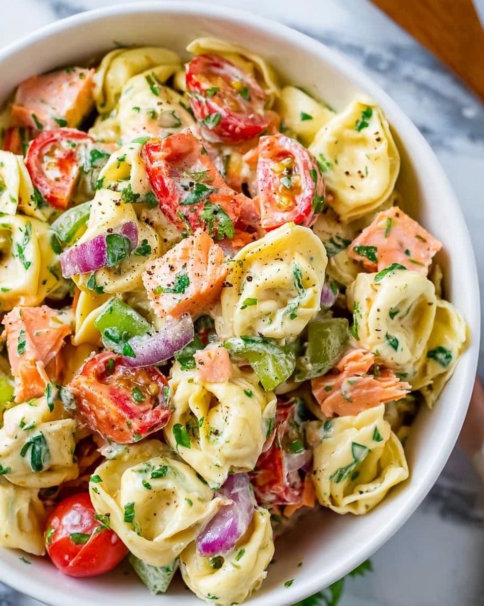A close-up of a white bowl filled with a colorful tortellini salad showing three main layers. The bottom layer is creamy yellow tortellini pasta with a smooth texture, scattered evenly throughout the bowl. On top of the pasta, there are chopped pieces of red and green bell peppers, along with halved bright red cherry tomatoes, giving a fresh and vibrant look. Mixed within these layers are chunks of pinkish-orange salmon and small bits of purple onion for contrast. The salad is lightly coated in a creamy dressing with some green leafy herbs sprinkled over the top, adding freshness and texture. The bowl sits on a surface with a white marbled texture. Photo taken with an iphone --ar 4:5 --v 7