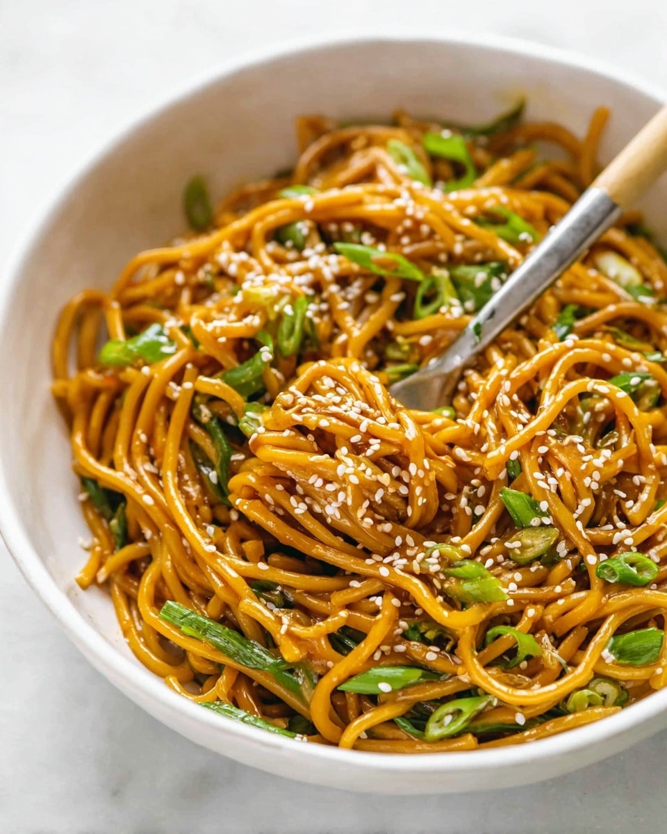 A white bowl filled with a generous serving of orange-brown noodles mixed with green chopped scallions, topped with white sesame seeds. The noodles appear saucy and glossy, with some sauce splattered on the inside rim of the bowl. A silver fork with a light wooden handle is partially stuck into the noodles, lifting some strands. The bowl sits on a white marbled surface. photo taken with an iphone --ar 4:5 --v 7