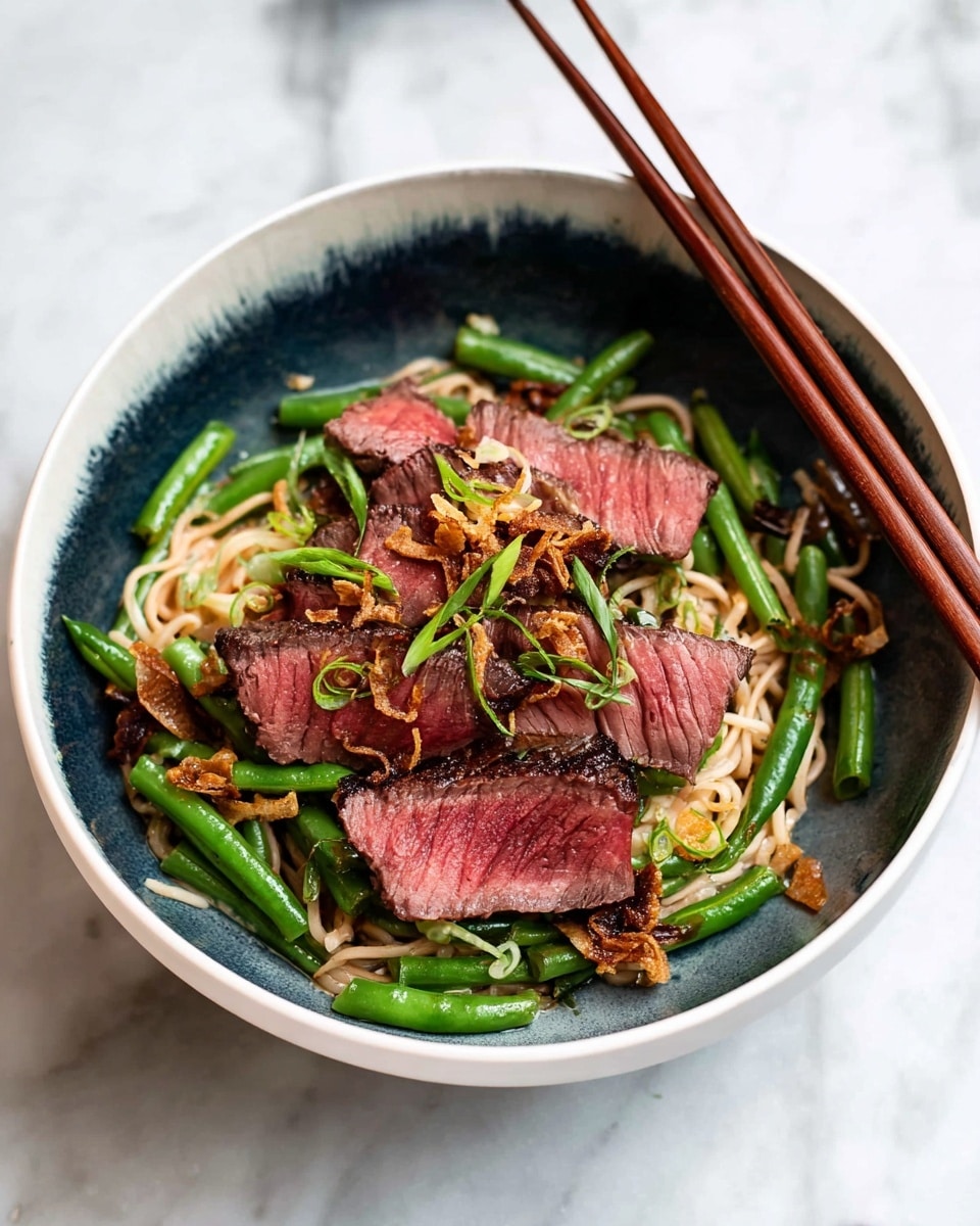 A bowl with a base layer of light beige noodles topped with bright green long beans scattered around. Over the beans, there are five slices of medium-rare steak with a pink center and a slightly charred, dark brown edge arranged in the middle. The steak is garnished with small rings of fresh green onions and thin, crispy fried shallots scattered on top. Two wooden chopsticks rest diagonally on the right side of the bowl. The bowl itself is white with a dark blue inner gradient, set on a white marbled surface. Photo taken with an iphone --ar 4:5 --v 7