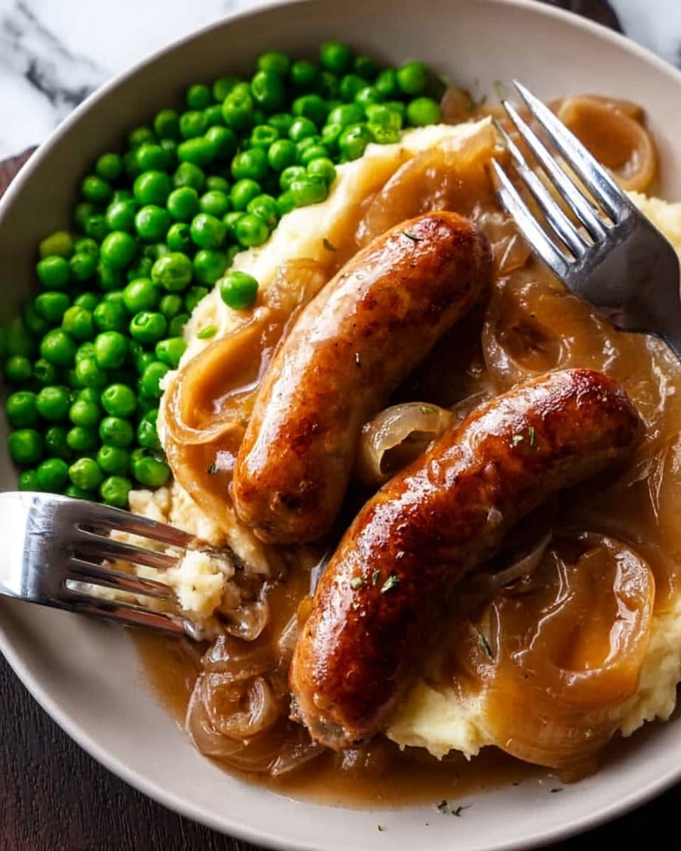 A white plate holds two browned sausages on top of a golden layer of mashed potatoes covered with a thick, light brown onion gravy with visible soft onion slices. To the left of the sausages, there is a bright green pile of cooked peas. A fork is placed on the right edge of the plate, partially touching a sausage, and a knife is on the left side, resting on the plate near the peas. The background is a white marbled texture. photo taken with an iphone --ar 4:5 --v 7