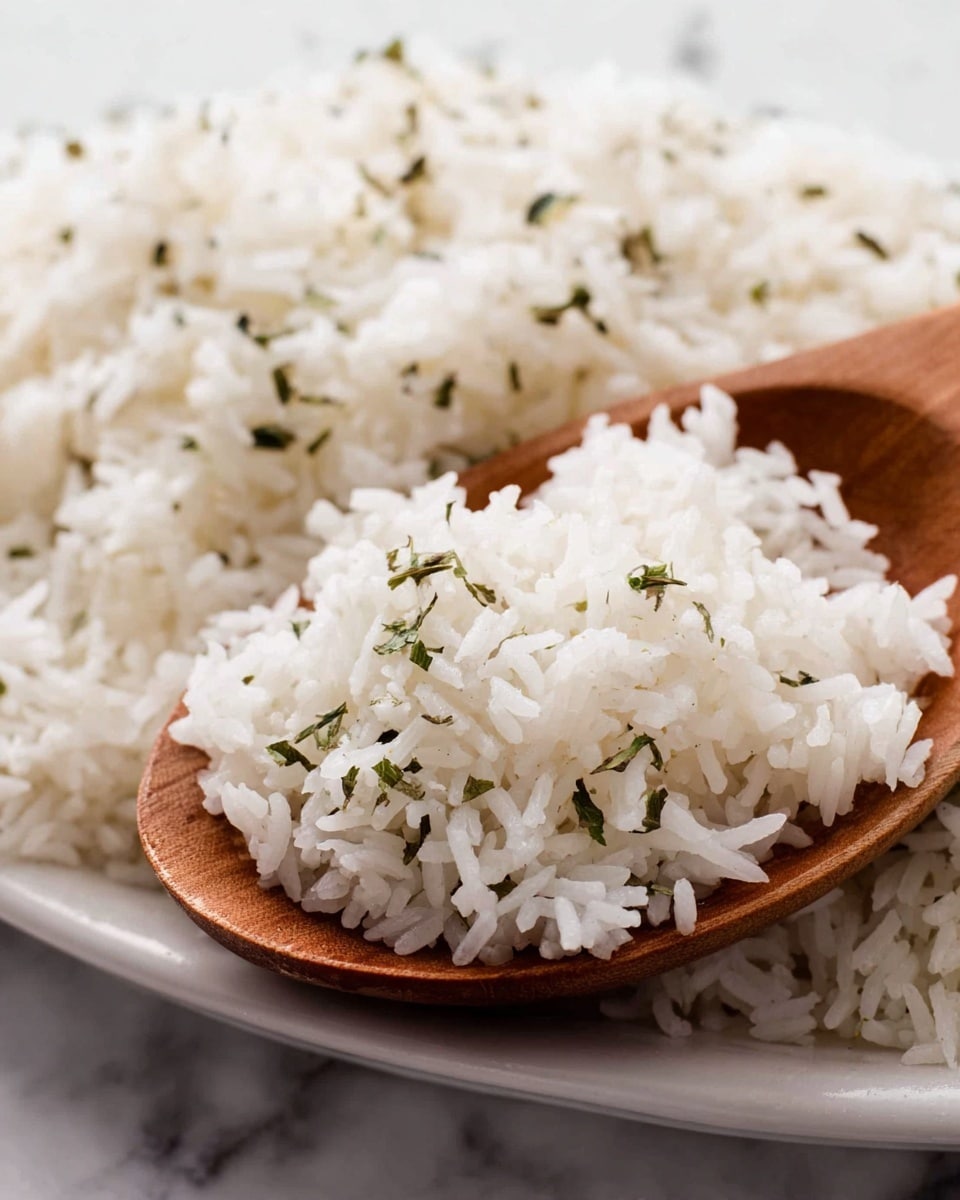 A close-up view of fluffy white rice with small green herb pieces scattered throughout, showing a soft texture with each grain distinct. The rice is heaped spooned slightly above the edge of a wooden spoon resting on a white plate, set against a white marbled surface. The composition focuses on the fine details of the rice grains and the warm tone of the wooden spoon. photo taken with an iphone --ar 4:5 --v 7