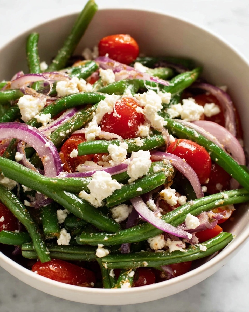 A close-up view of a fresh salad in a white bowl sitting on a white marbled surface, showing three main layers: the bottom layer has thin red onion slices with a shiny texture, the middle layer is made of bright green green beans cut into pieces, and the top layer features halved red cherry tomatoes with a glossy look and scattered white crumbled cheese, all mixed gently and lightly coated with dressing, with some dressing visible in small pools on the tomato surfaces. photo taken with an iphone --ar 4:5 --v 7