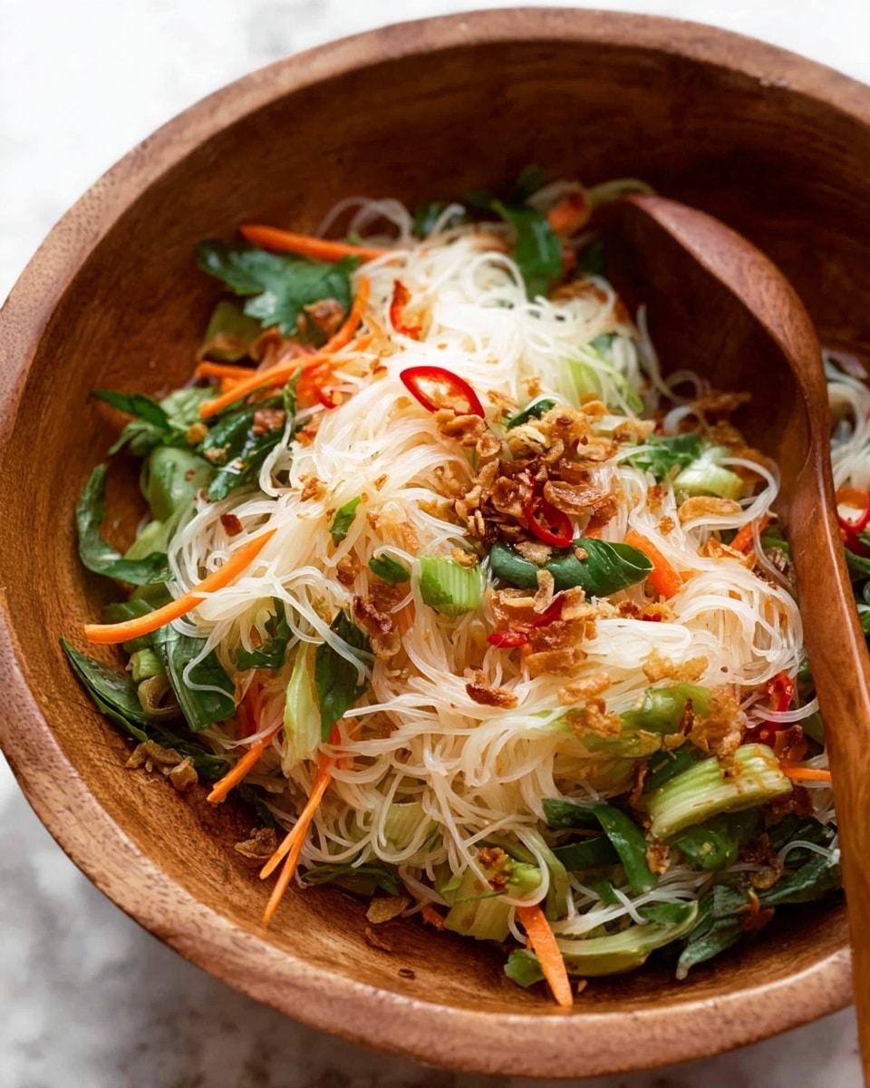 A close-up view of a wooden bowl filled with a colorful noodle salad, showing thin white rice vermicelli noodles as the main base mixed with thin orange carrot strips, small light green scallion rings, and bright red chili slices. Scattered dark brown crispy fried shallots or garlic pieces add texture on top, while fresh green cilantro leaves and other leafy greens are spread throughout. A wooden spoon is partly visible resting inside the bowl. The background features a white marbled surface. photo taken with an iphone --ar 4:5 --v 7