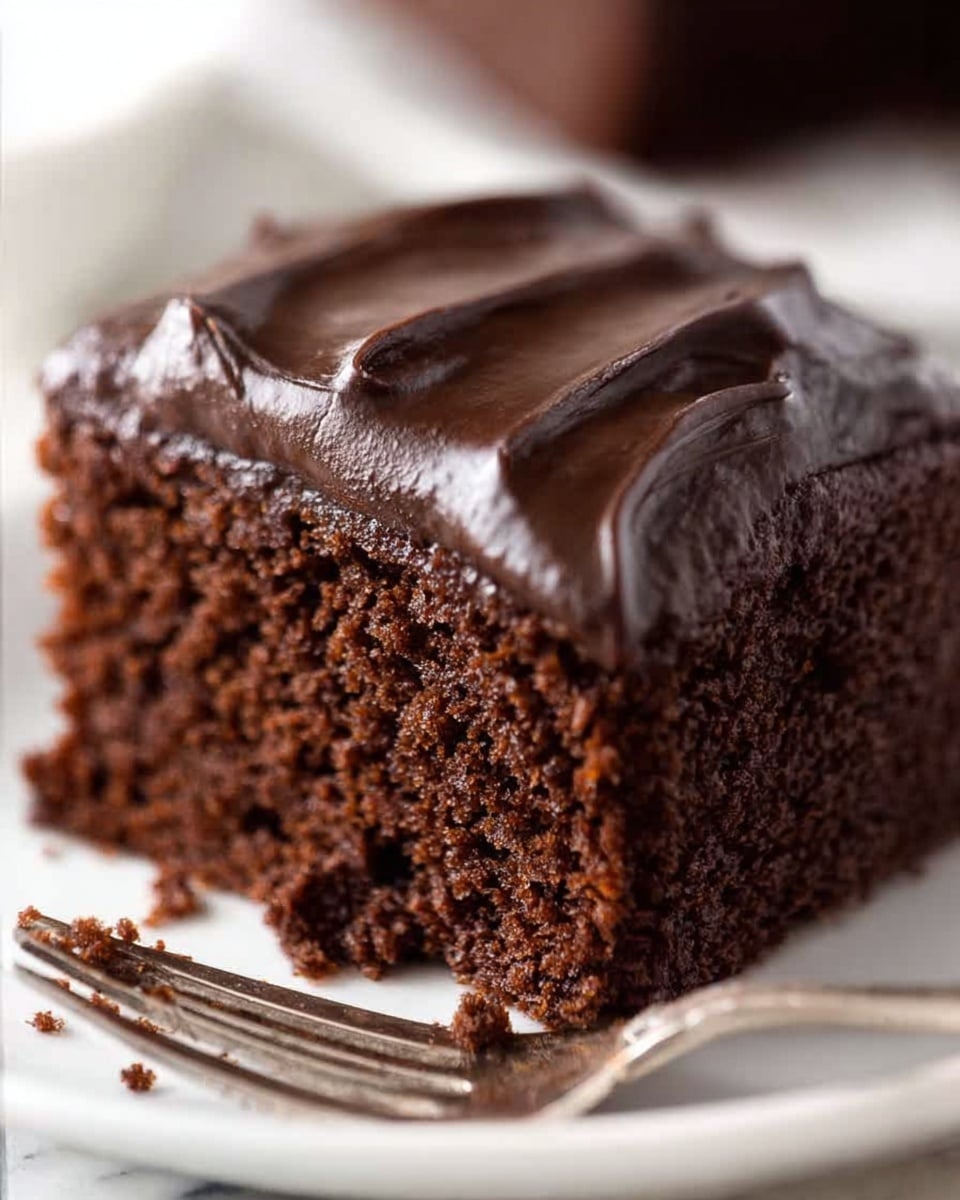 A close-up view of a single slice of moist chocolate cake with one thick layer. The cake layer is dark brown and soft with a slightly crumbly texture. On top, there is a thick, smooth layer of glossy dark chocolate frosting with visible swirled ridges. The slice sits on a white plate placed on a white marbled surface. Cake crumbs are scattered around the plate near a metal fork in the background. photo taken with an iphone --ar 4:5 --v 7