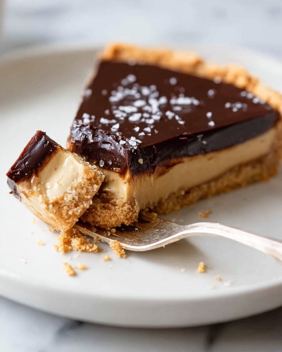 A slice of dessert with three clear layers sits on a white plate over a white marbled texture. The bottom layer is a crumbly light brown crust with a rough texture. The middle layer is a smooth, creamy peanut butter-colored filling, thick and soft. The top layer is a shiny, dark chocolate glaze, smooth and glossy, sprinkled with small flakes of white sea salt. A spoon is pulling a bite-sized piece from the slice, showing the layers clearly. photo taken with an iphone --ar 4:5 --v 7